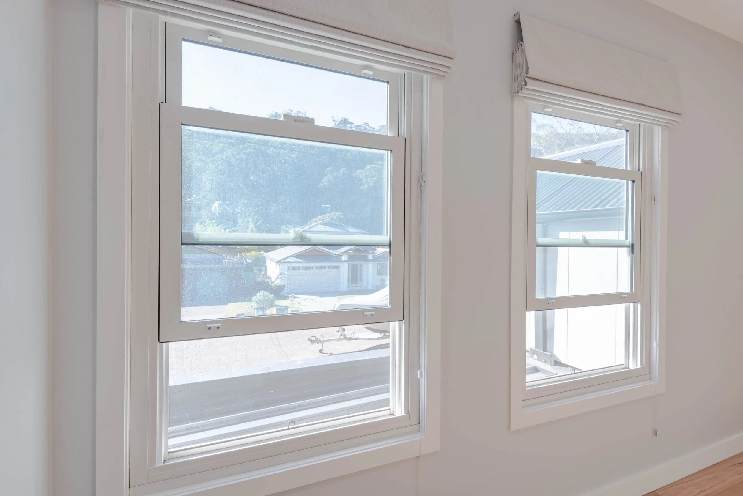 Two double-hung windows with white trim and beige window valances in a white-walled room, with a view of houses and trees outside.