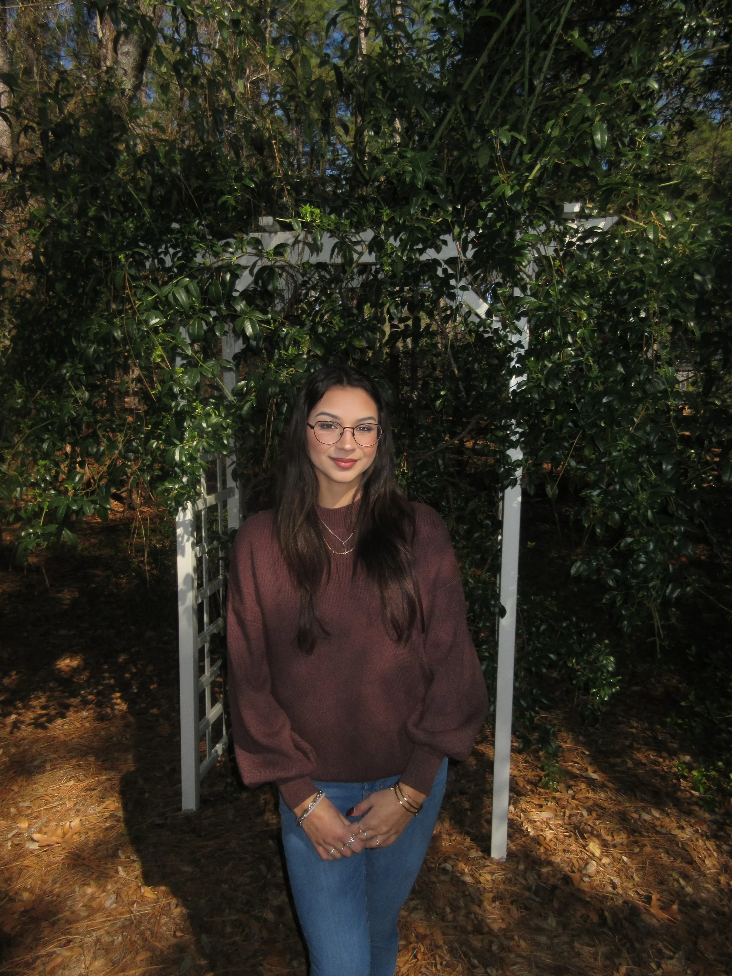 A young woman with long dark hair, glasses, and a brown sweater standing outdoors in front of greenery and a white garden arch.