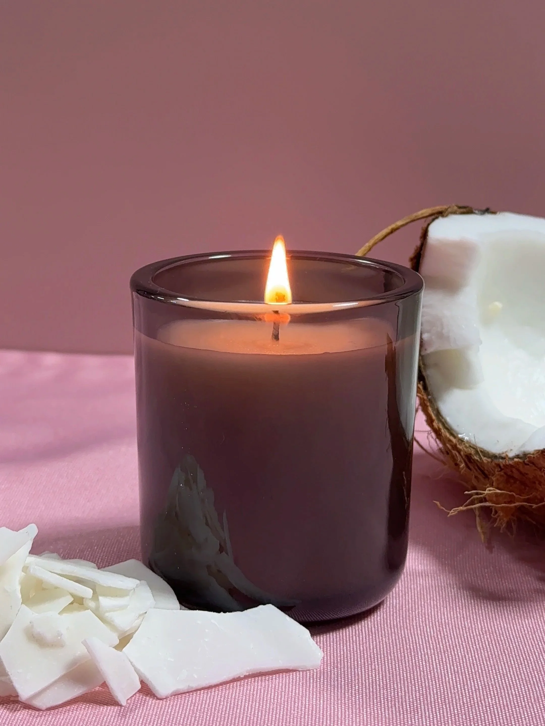 A lit candle in a gray glass holder, with coconut pieces and half a coconut on a pink surface. skincare Bay Area facial candle soap organic skincare routine bath and body Sonoma lux living lux unfiltered coastal living luxurious