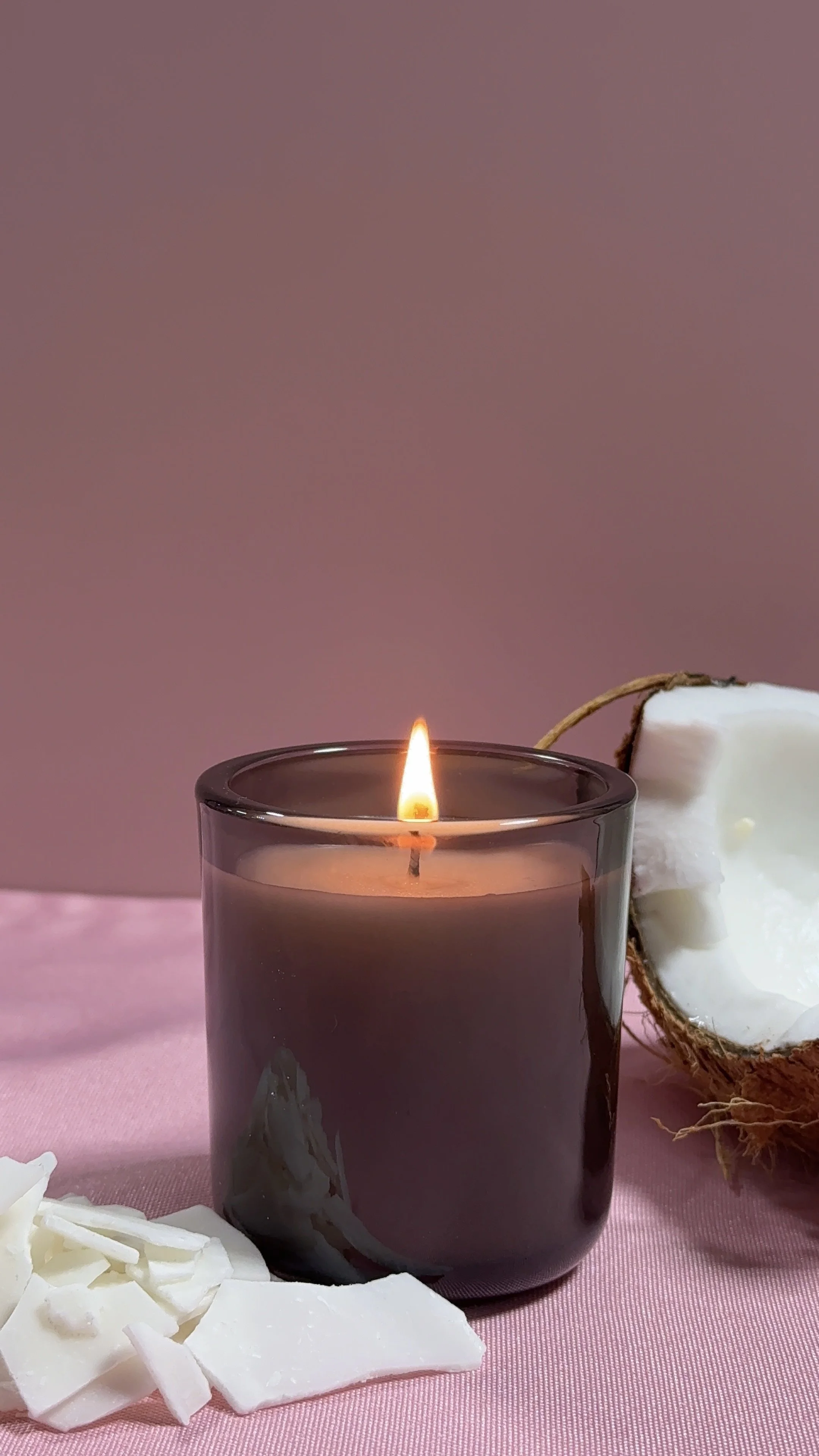 A lit brown candle in a glass holder on a pink surface next to a halved coconut and white coconut flakes.