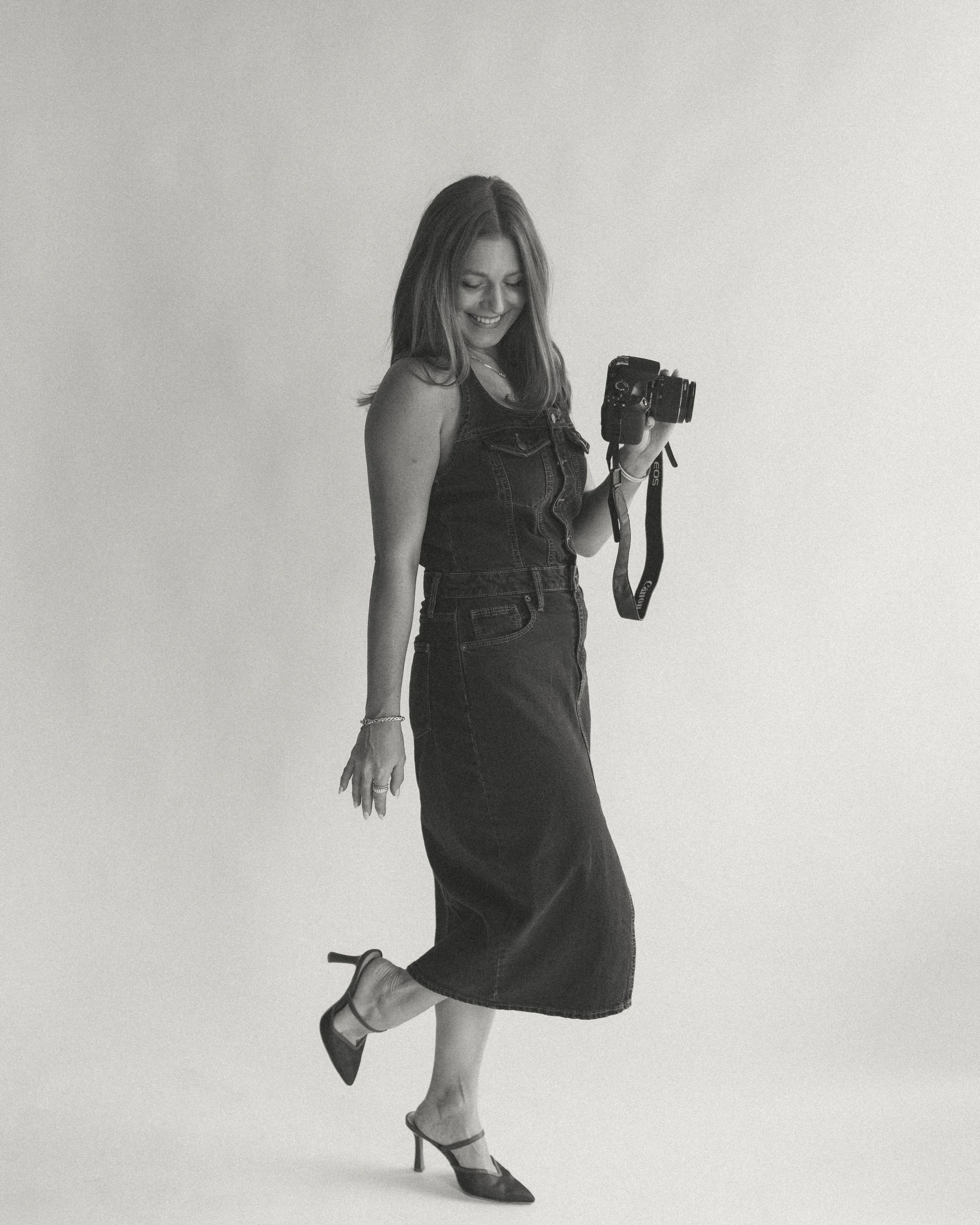 A woman in a denim dress and high heels holding a camera, smiling and looking down, against a plain background. media sharp agency owner ceo women.