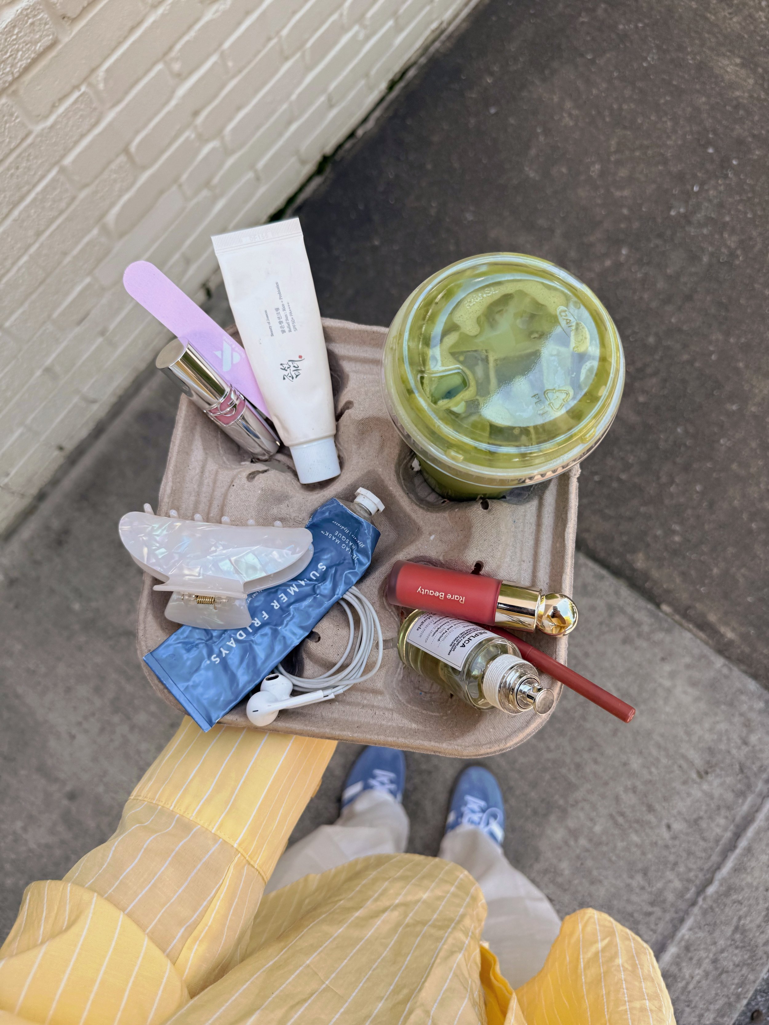 Person standing on sidewalk holding a cardboard tray with various personal care items, a large green smoothie in a clear cup, and ground in the background.