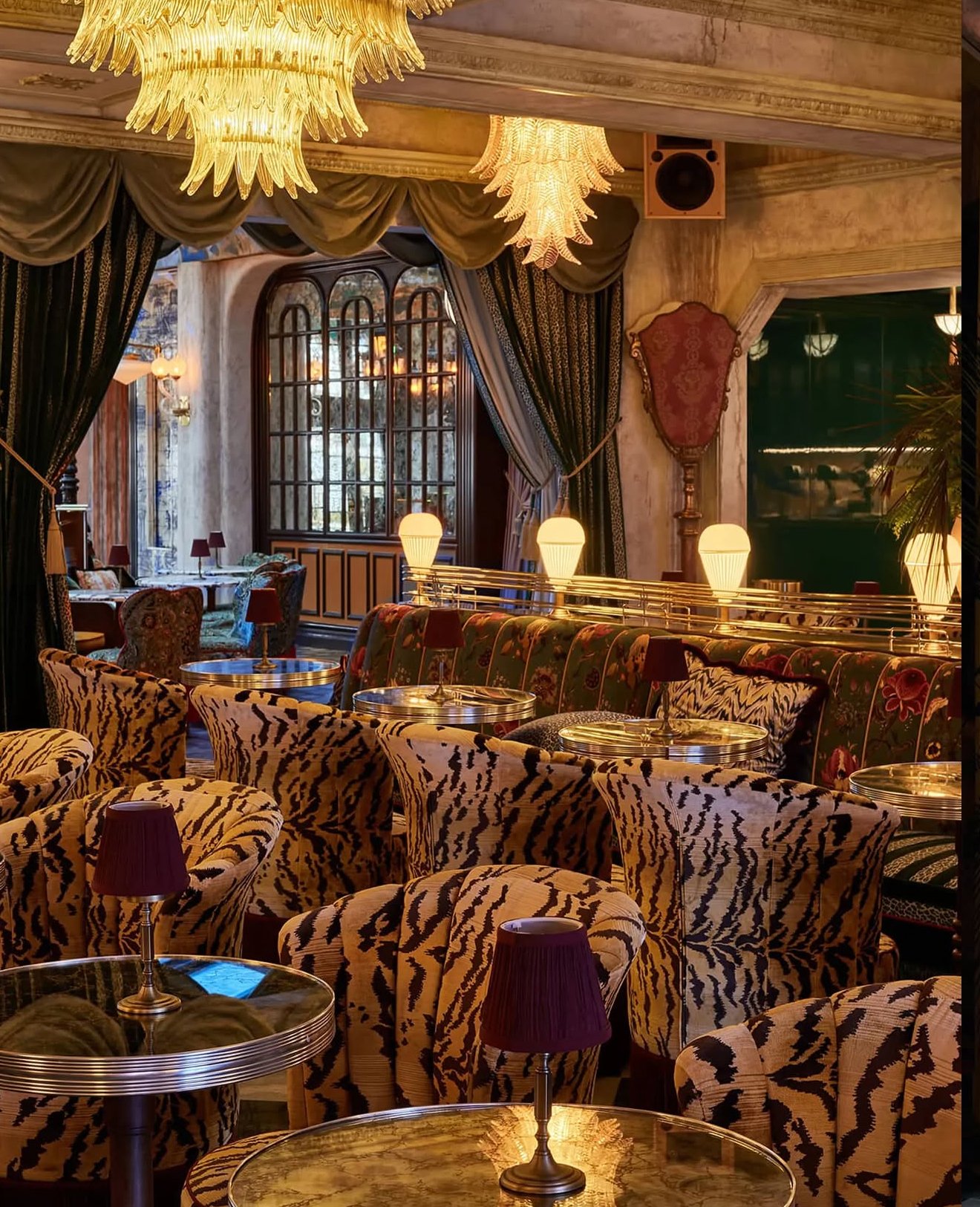 Luxurious vintage-style lounge with plush chairs in zebra print, small tables with purple lamps, ornate curtains, and golden chandeliers.