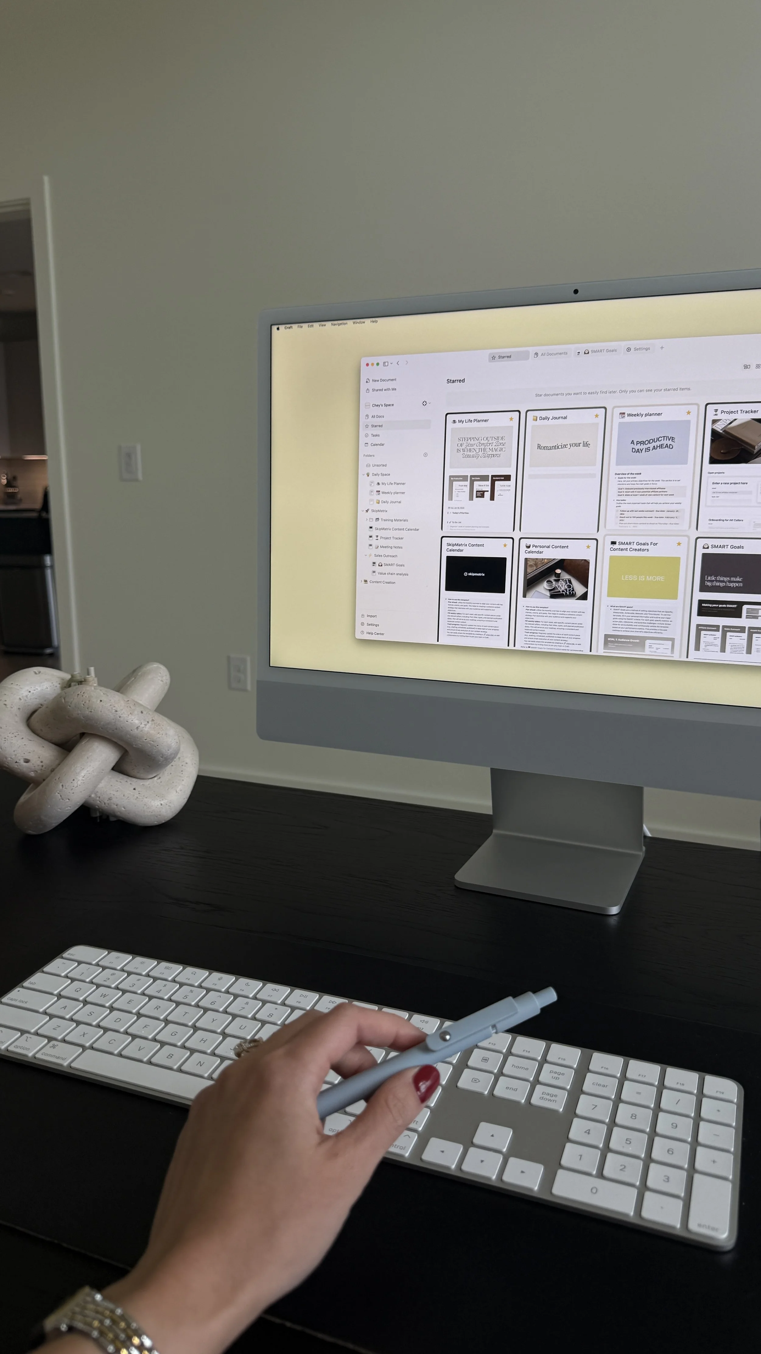 A person's hand with red nail polish holding a stylus near a white keyboard on a black desk. A computer monitor displays a digital planning or note-taking application and is in a room with a plain light-colored wall. There is a decorative sculpture on the desk resembling a chain link.