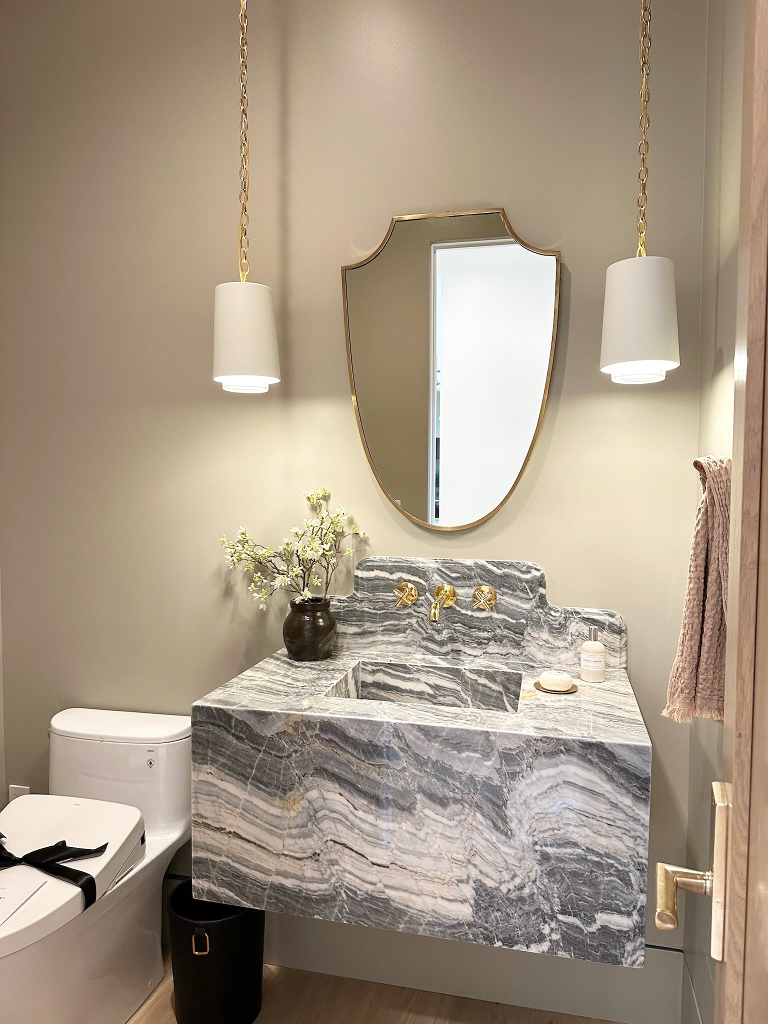 A modern bathroom featuring a marble sink with gold faucet fixtures, a mirror, a white toilet, and wall-mounted pendant lights.  small vase with flowers and toiletries are placed on the sink. Bay Area interior designer home remodel home decor realtor