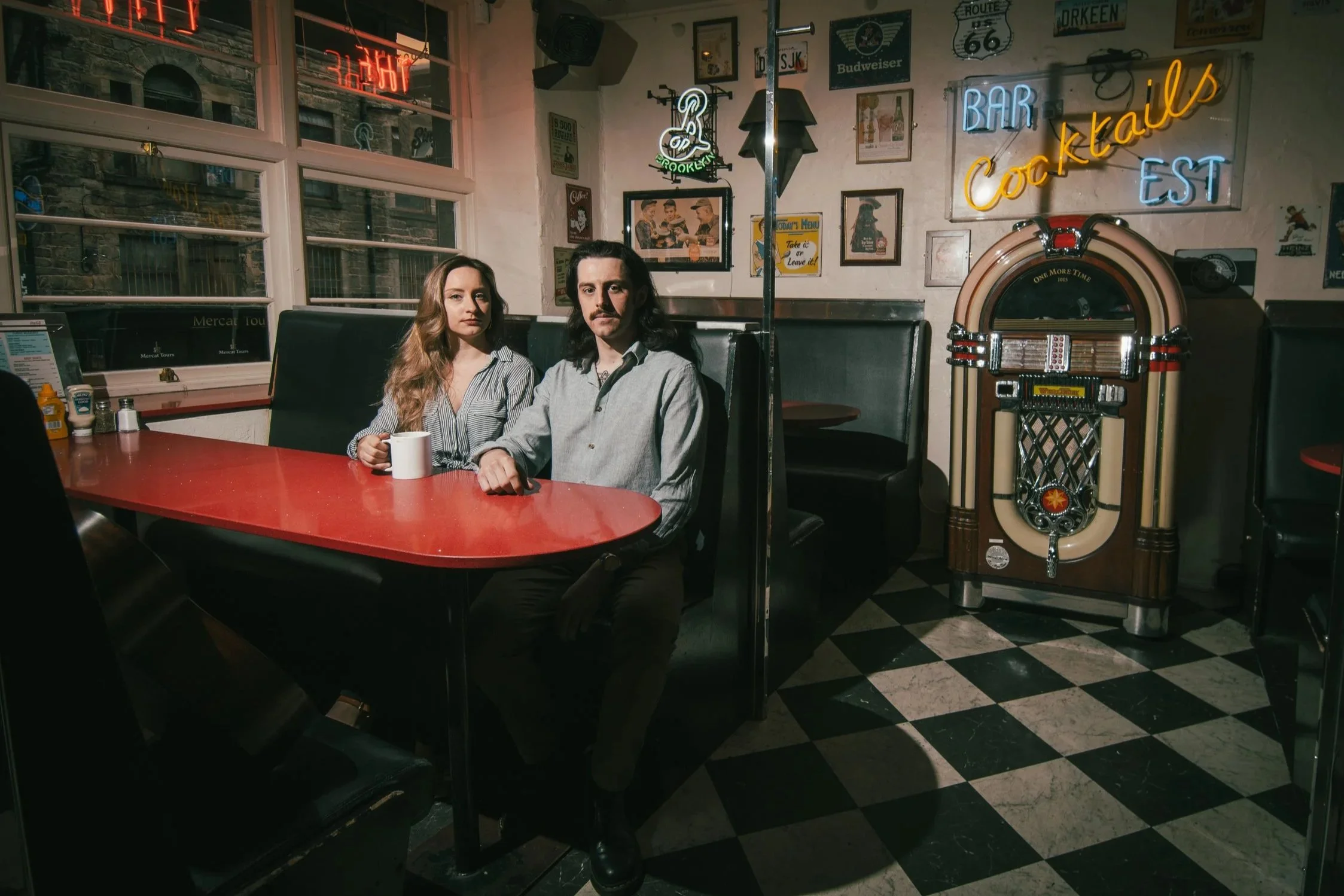 Honeycomb Acoustic Duo musicians sitting at a red table in a vintage-style bar or diner with neon signs that read "Bar" and "Cocktails" on the wall behind them, along with various framed pictures and a jukebox.