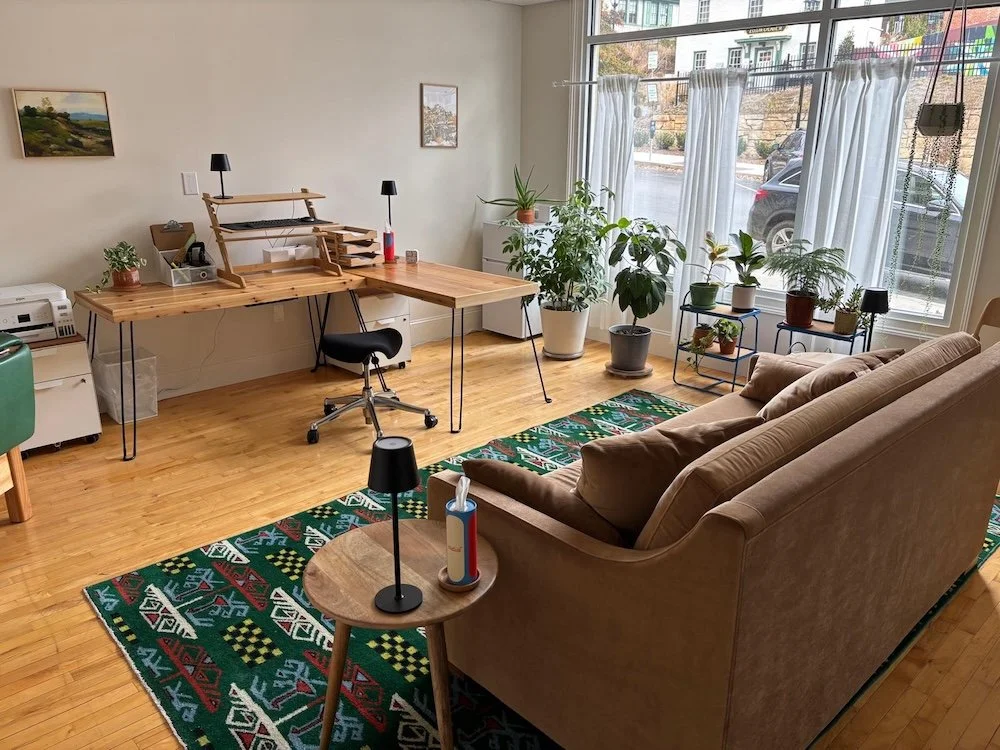 Living room with a brown sofa, colorful patterned rug, wooden desk with office items, and large window with white curtains and several potted plants outside.