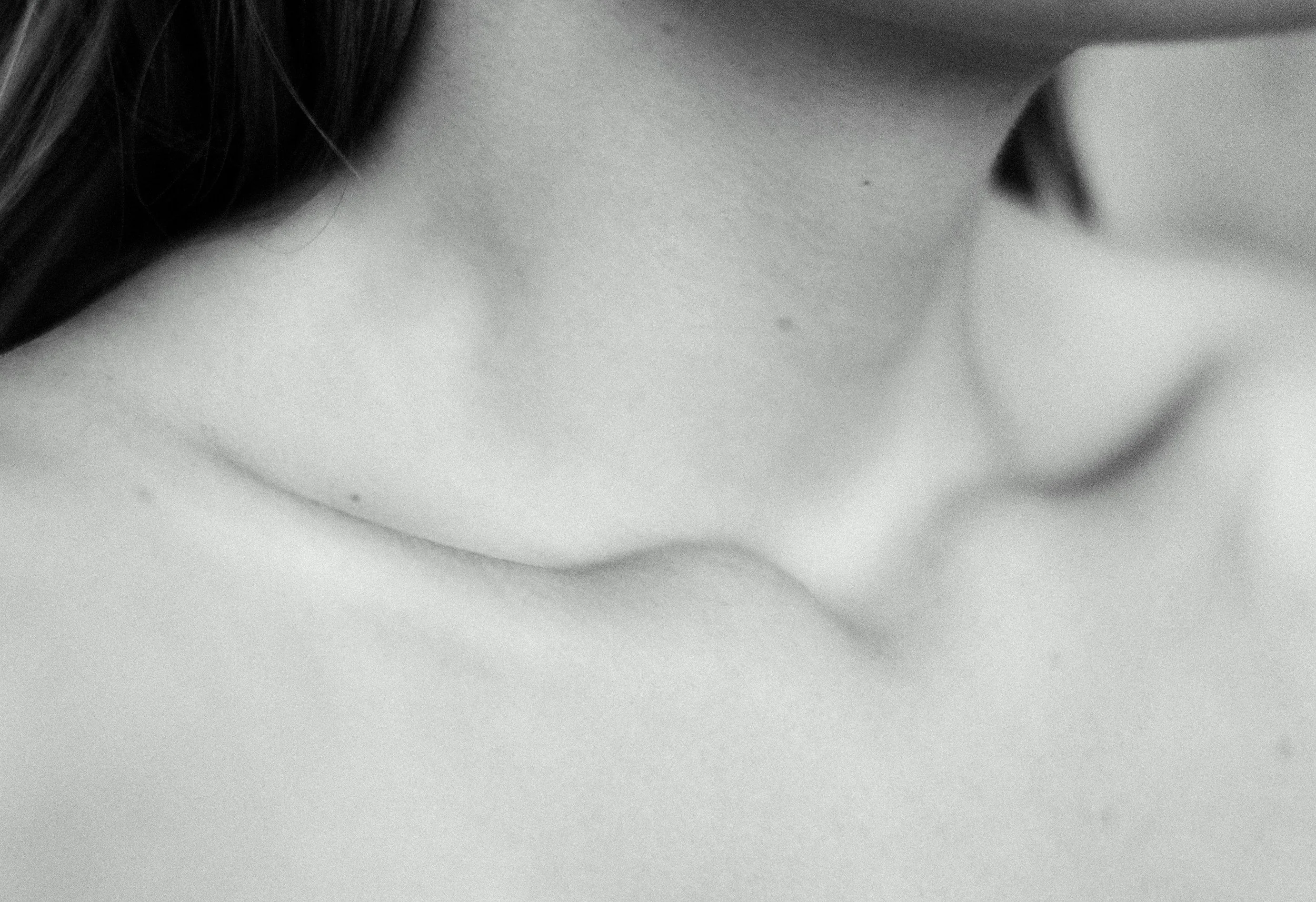 Close-up black and white photo of a person's neck and collarbone area, with part of their dark hair visible.
