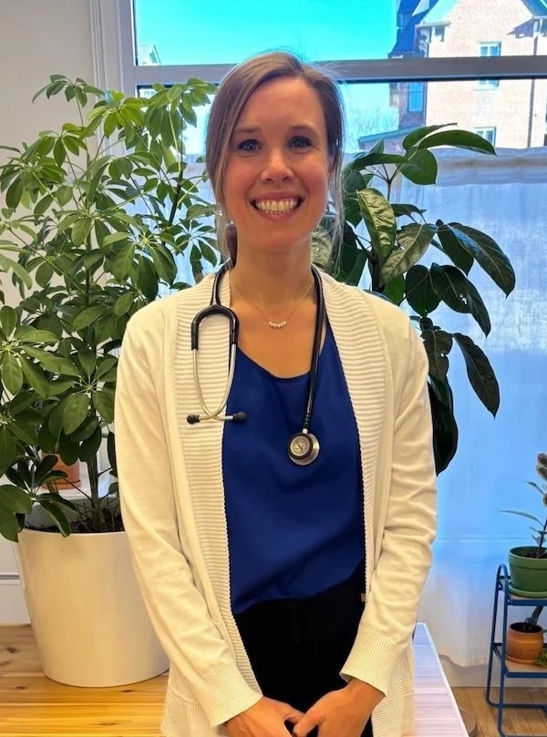 A smiling woman, likely a healthcare professional, standing indoors near green potted plants, wearing a stethoscope around her neck and a beige cardigan over a blue top, with a window showing buildings in the background.
