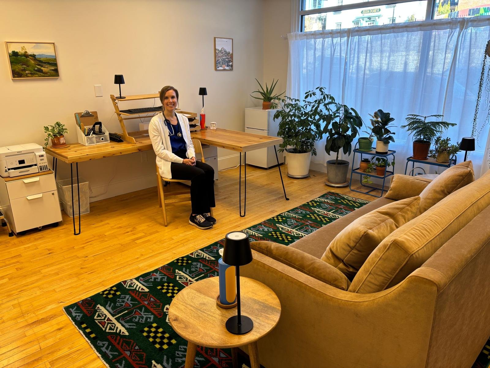 A woman in a white coat, sitting on a chair in a well-lit office with a wooden desk, leafy plants, and a beige sofa.