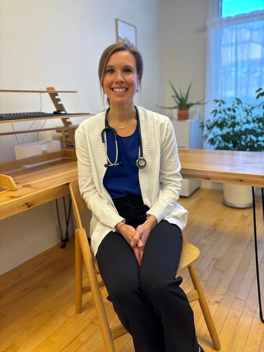A woman in medical scrubs with a stethoscope around her neck, sitting on a wooden chair in a room with wooden flooring, a wooden desk, and potted plants in the background.