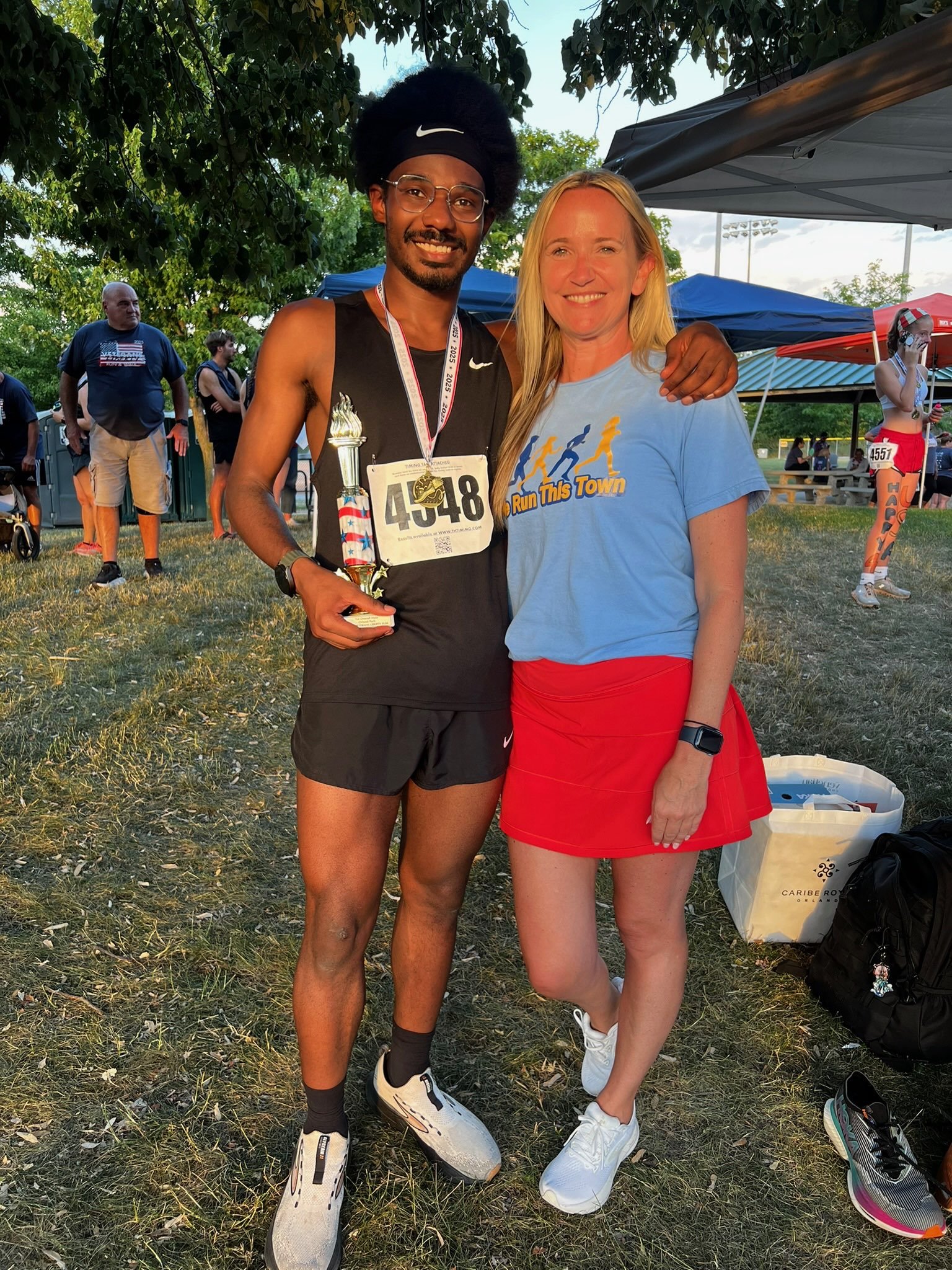 A young male runner with a medal, holding a trophy, standing next to a woman in a blue T-shirt and red skirt, both smiling at a track event.