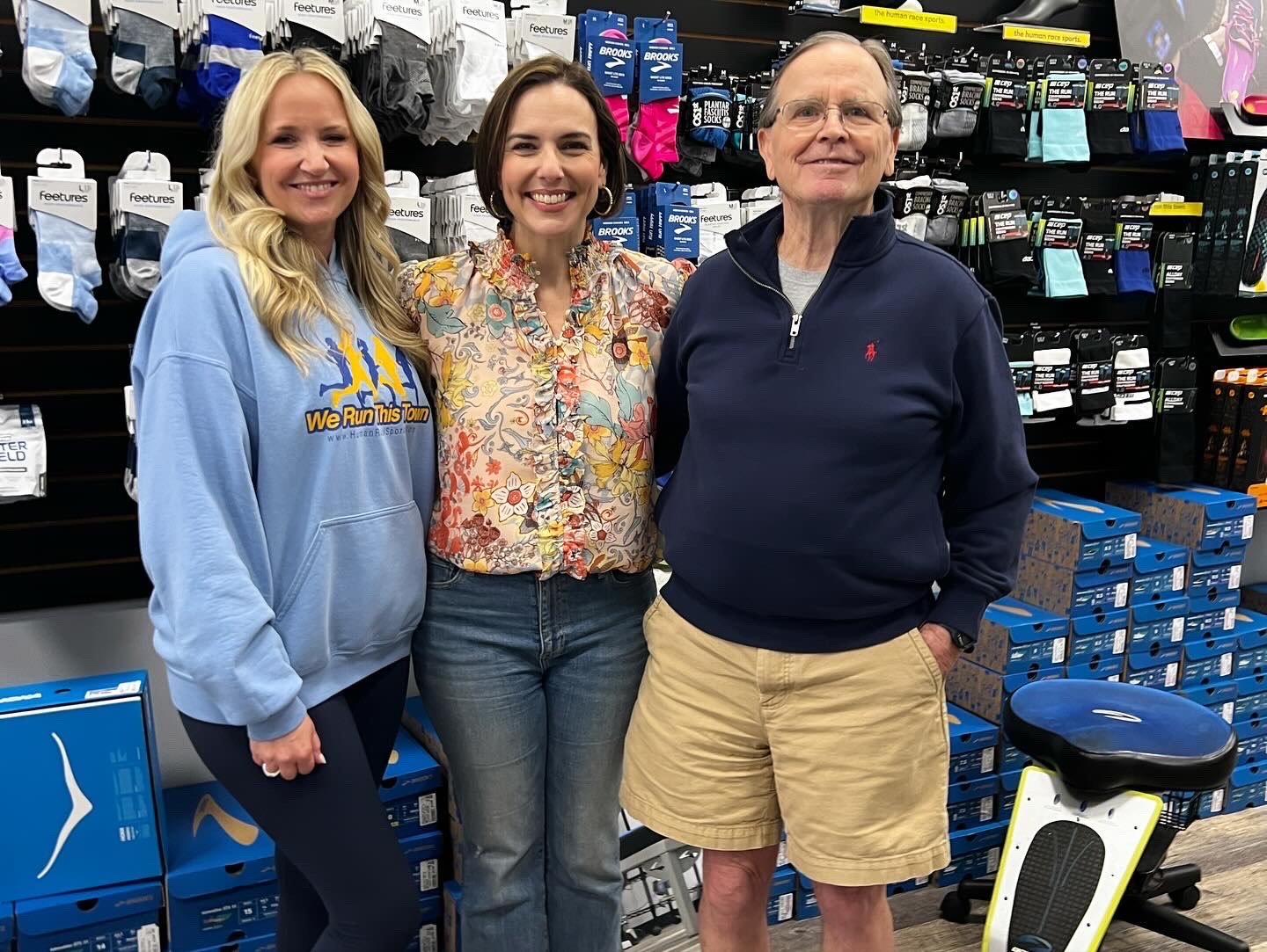 Three people standing inside a sporting goods store, surrounded by boxes of athletic shoes and socks, smiling at the camera.