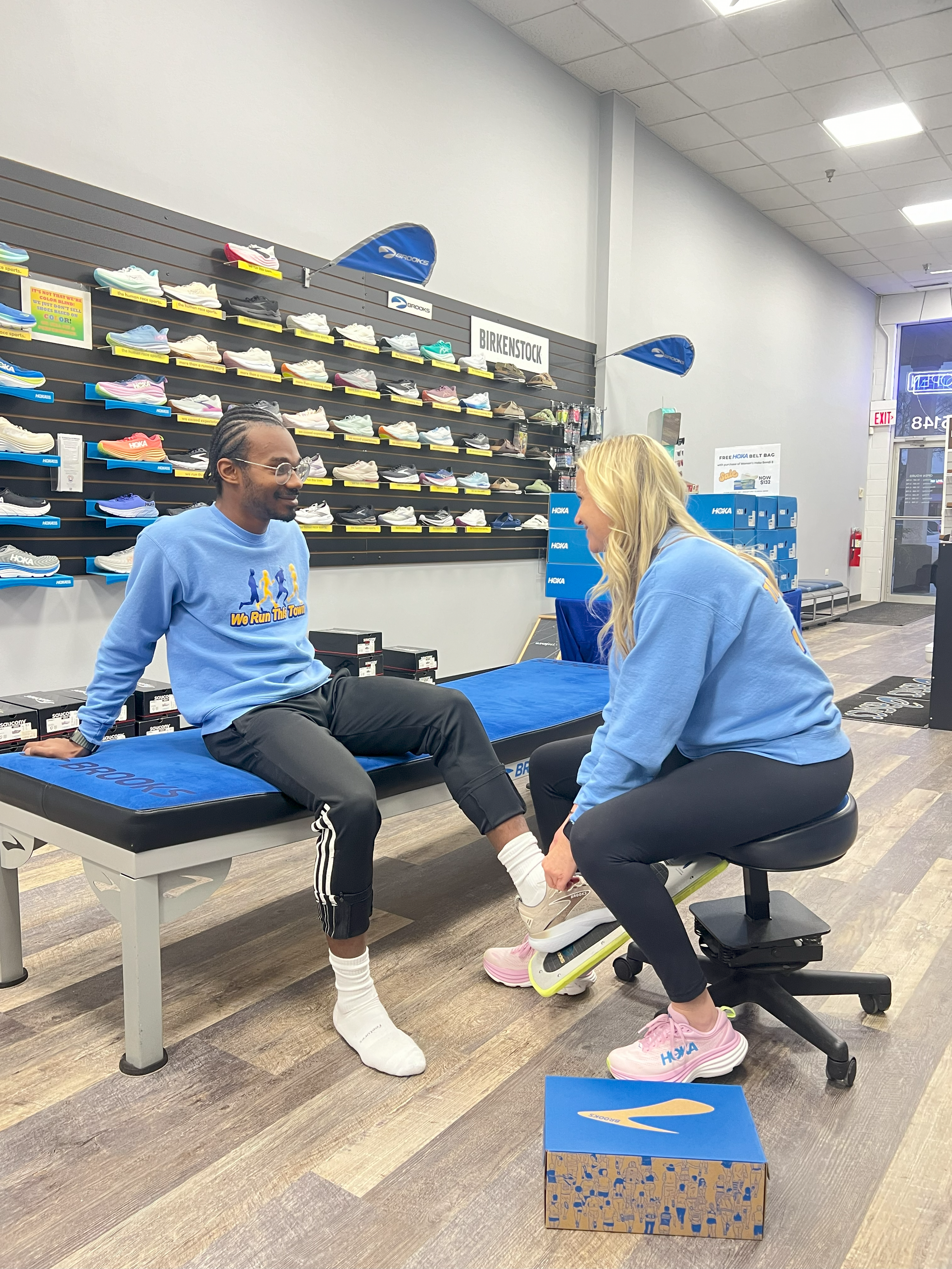 A woman helps a man try on running shoes inside a shoe store, with shelves of sneakers displayed behind them.