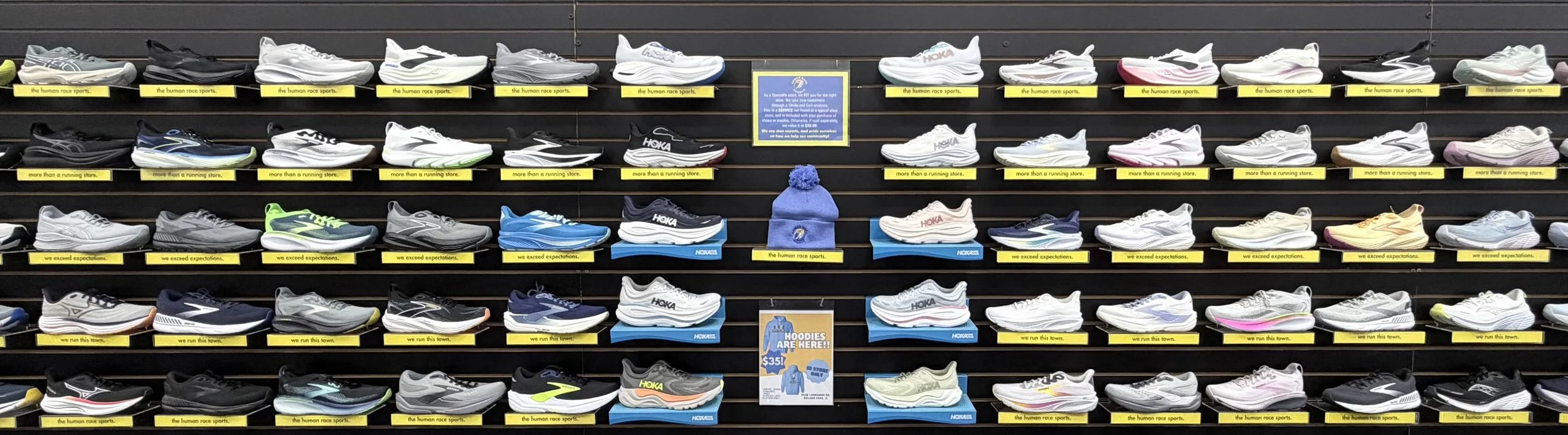 Wall display of various athletic shoes organized on black shelves with yellow labels. The shoes are mostly white, black, gray, and blue, with some in other colors. There is a framed sign in the center and promotional posters below.