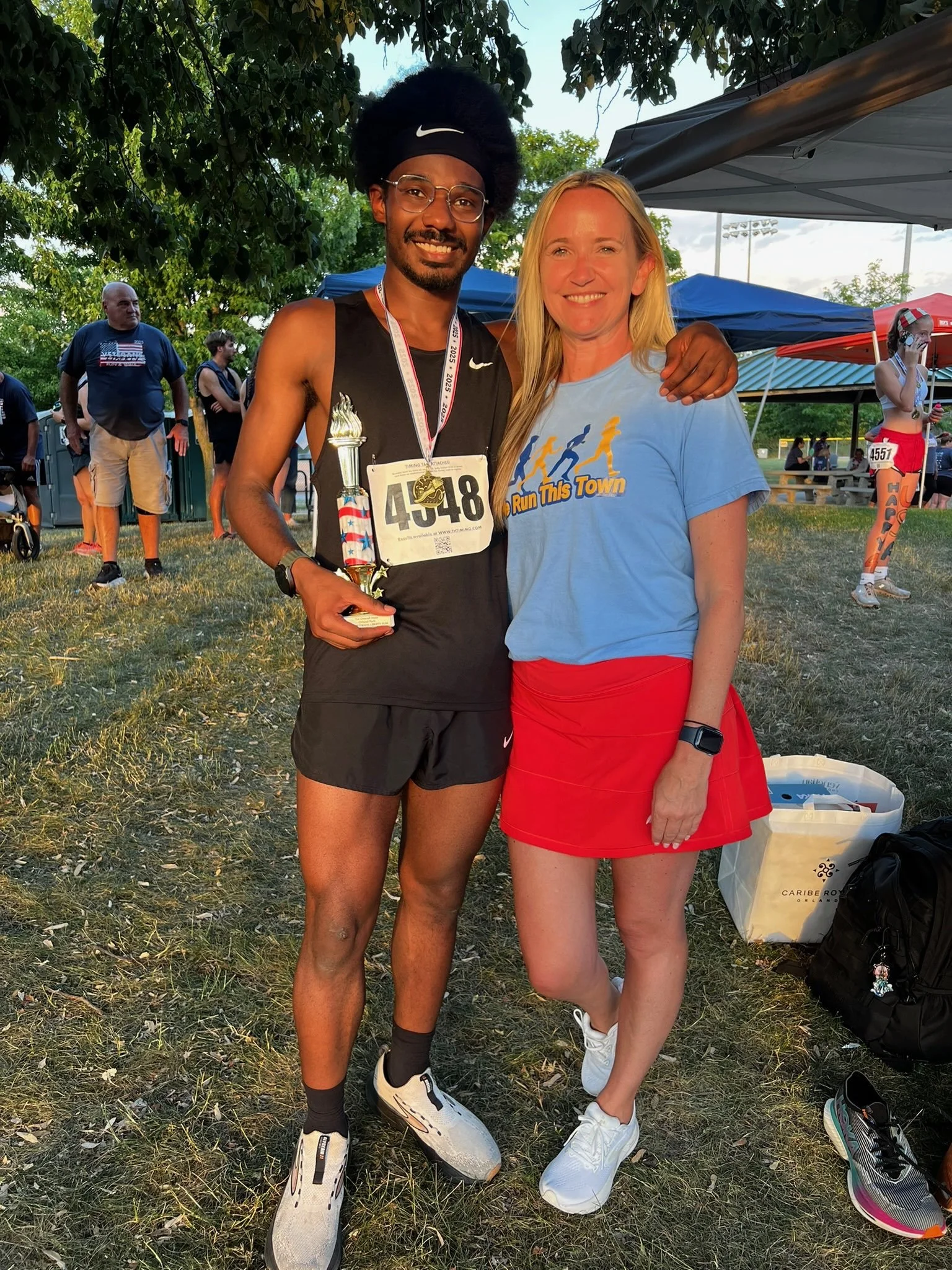 A man in running attire holding a trophy and wearing a medal, standing next to a woman in a blue T-shirt and red skirt, both smiling at an outdoor event with tents and other runners in the background.