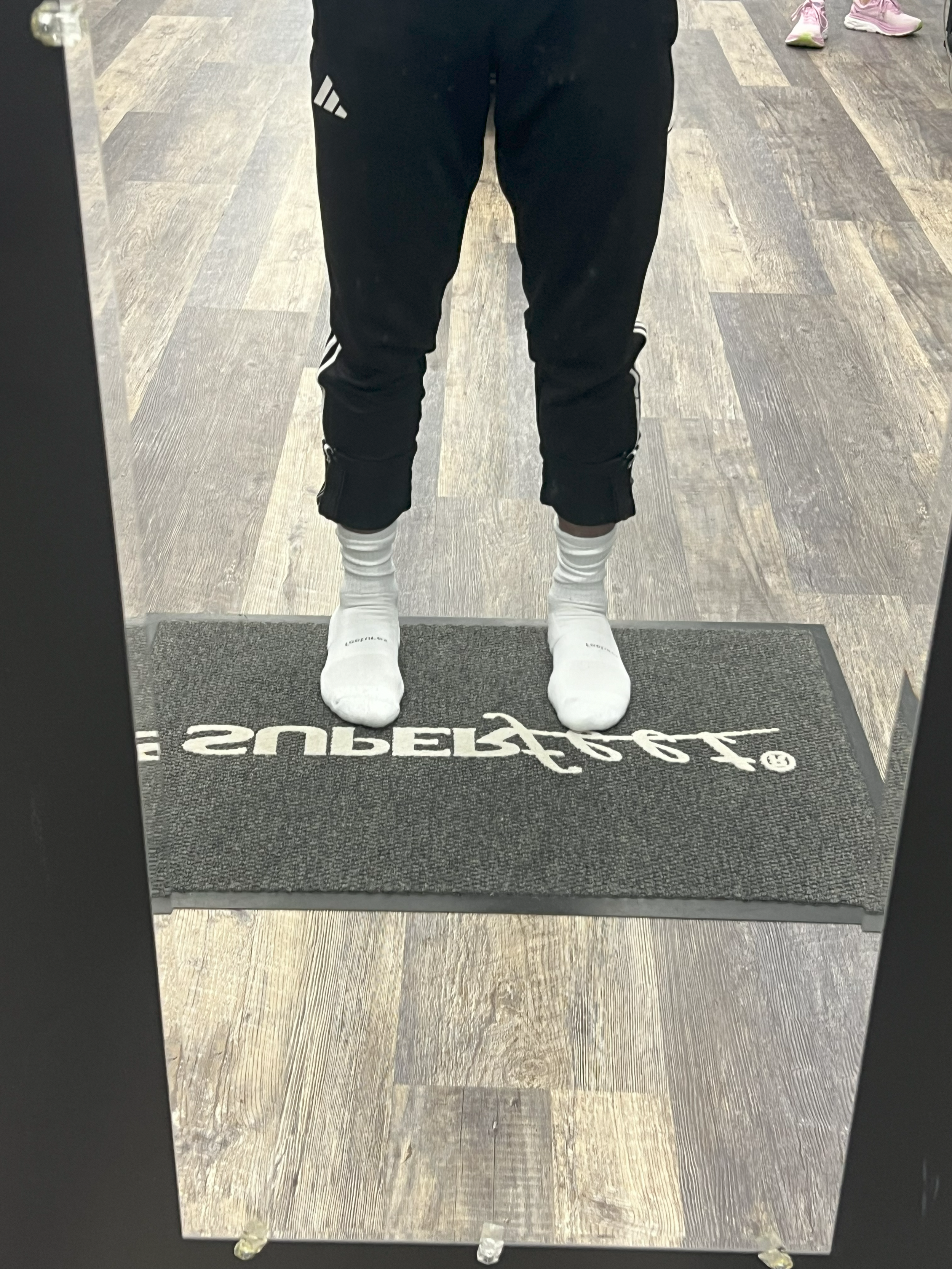 A person standing in front of a mirror taking a photo of their legs. They are wearing black athletic pants, white athletic socks, and white sneakers. The floor is wooden, and a black rug with white text is in front of the mirror.