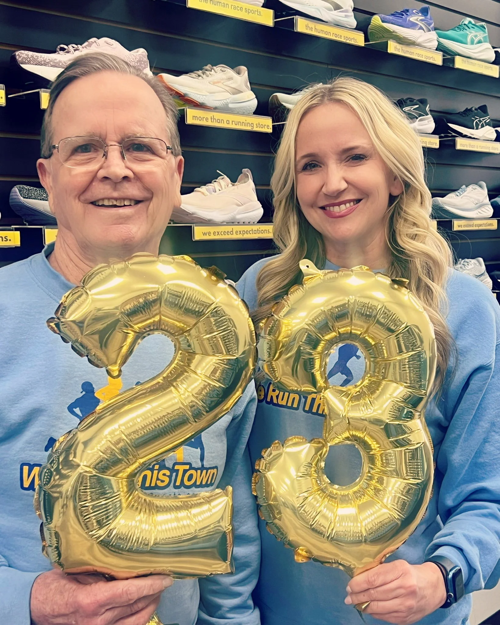 A man and woman smiling and holding gold balloons shaped as the numbers 2 and 3 in a sporting goods store, with shelves of running shoes in the background.
