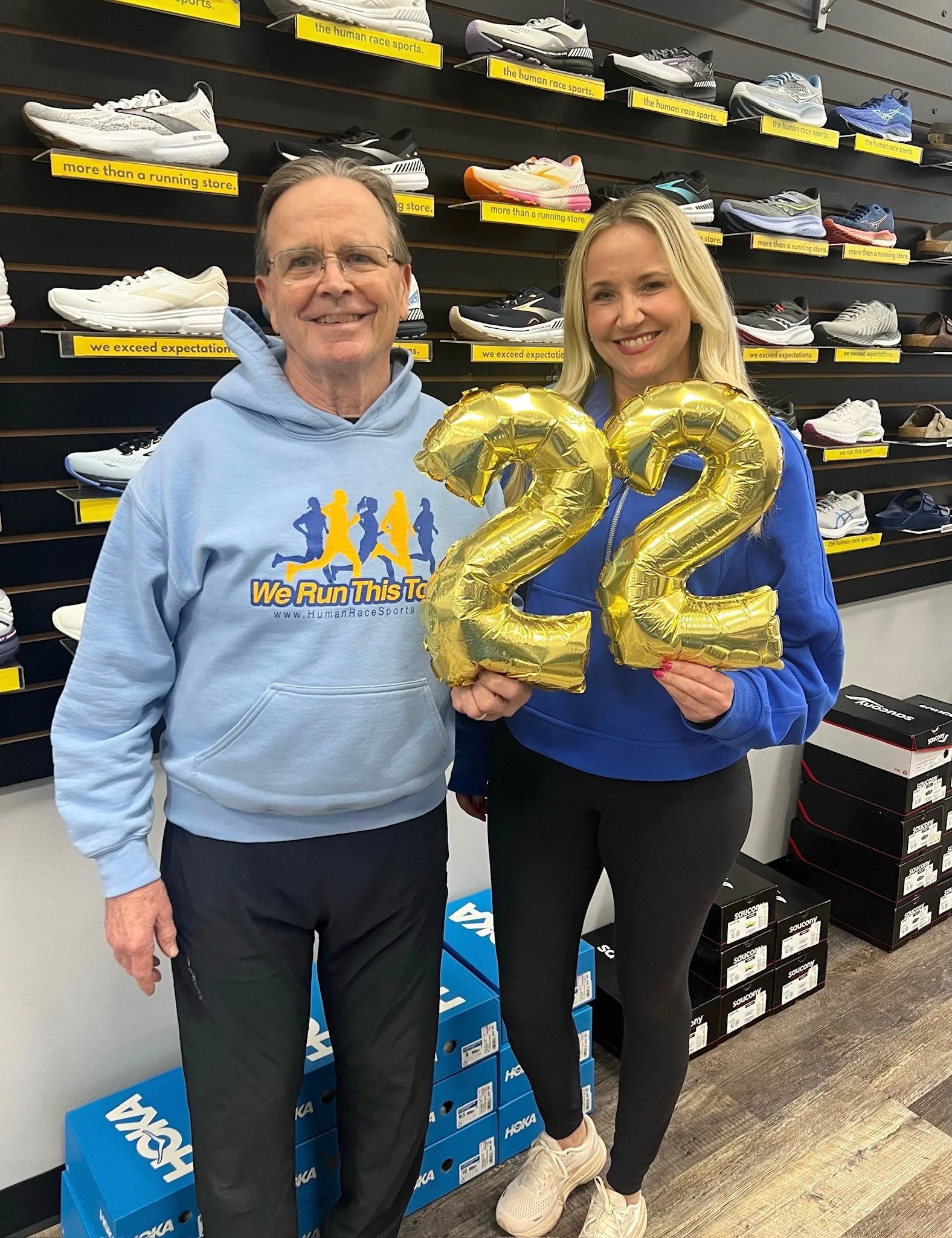 Two people standing in a shoe store, holding gold balloon numbers '22'. The man is wearing glasses, a light blue hoodie with 'We Run This Town' logo, and black pants. The woman has long blonde hair, a blue jacket, black leggings, and sneakers. They a