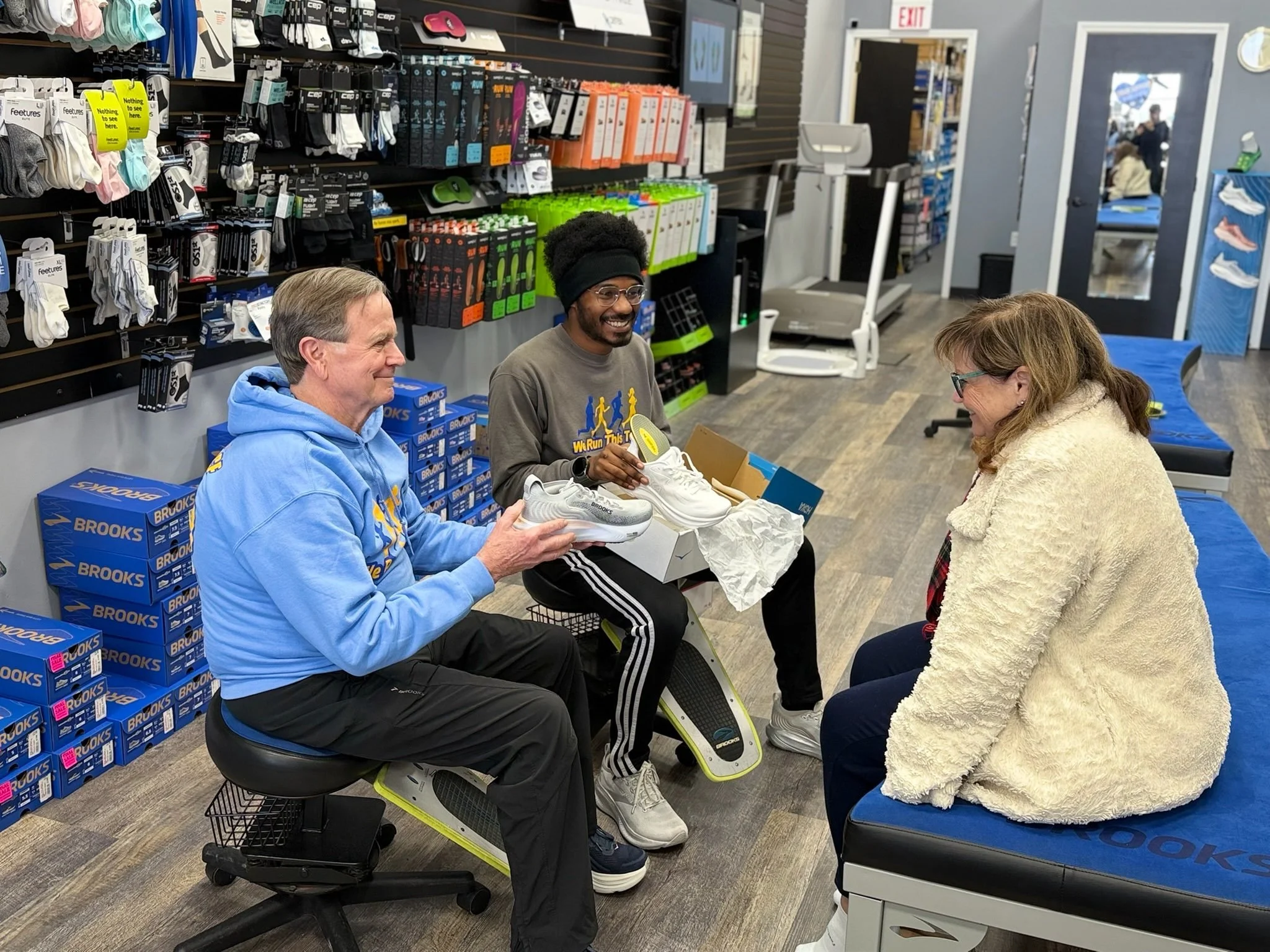 Three people are seated in a shoe store; two men are sitting on stools, holding white running shoes, and a woman is sitting on a bench across from them, smiling and engaging in conversation.