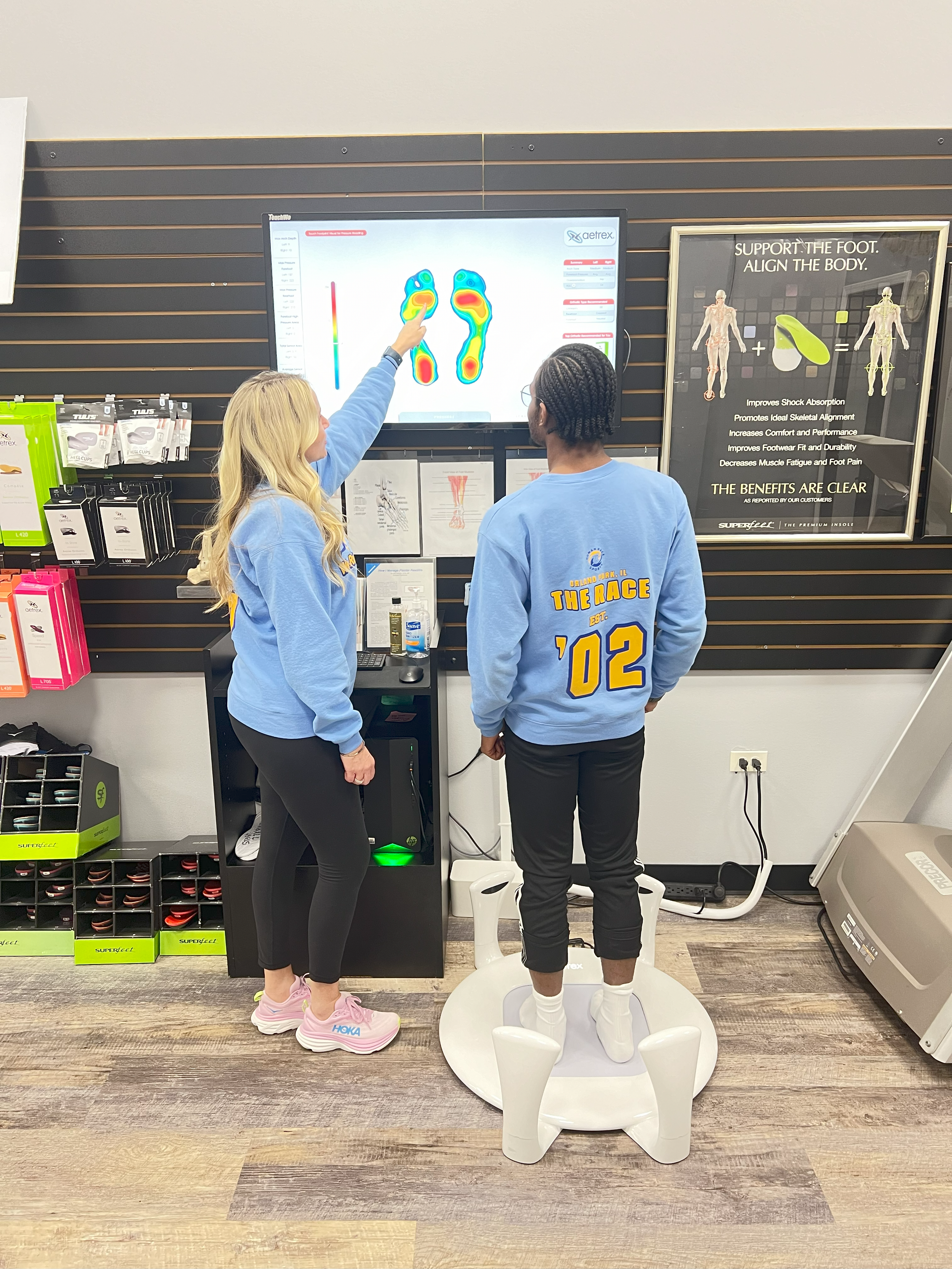 A woman is pointing at a foot pressure analysis on a large screen while a man stands on a pressure plate in a shoe store, receiving a foot analysis.