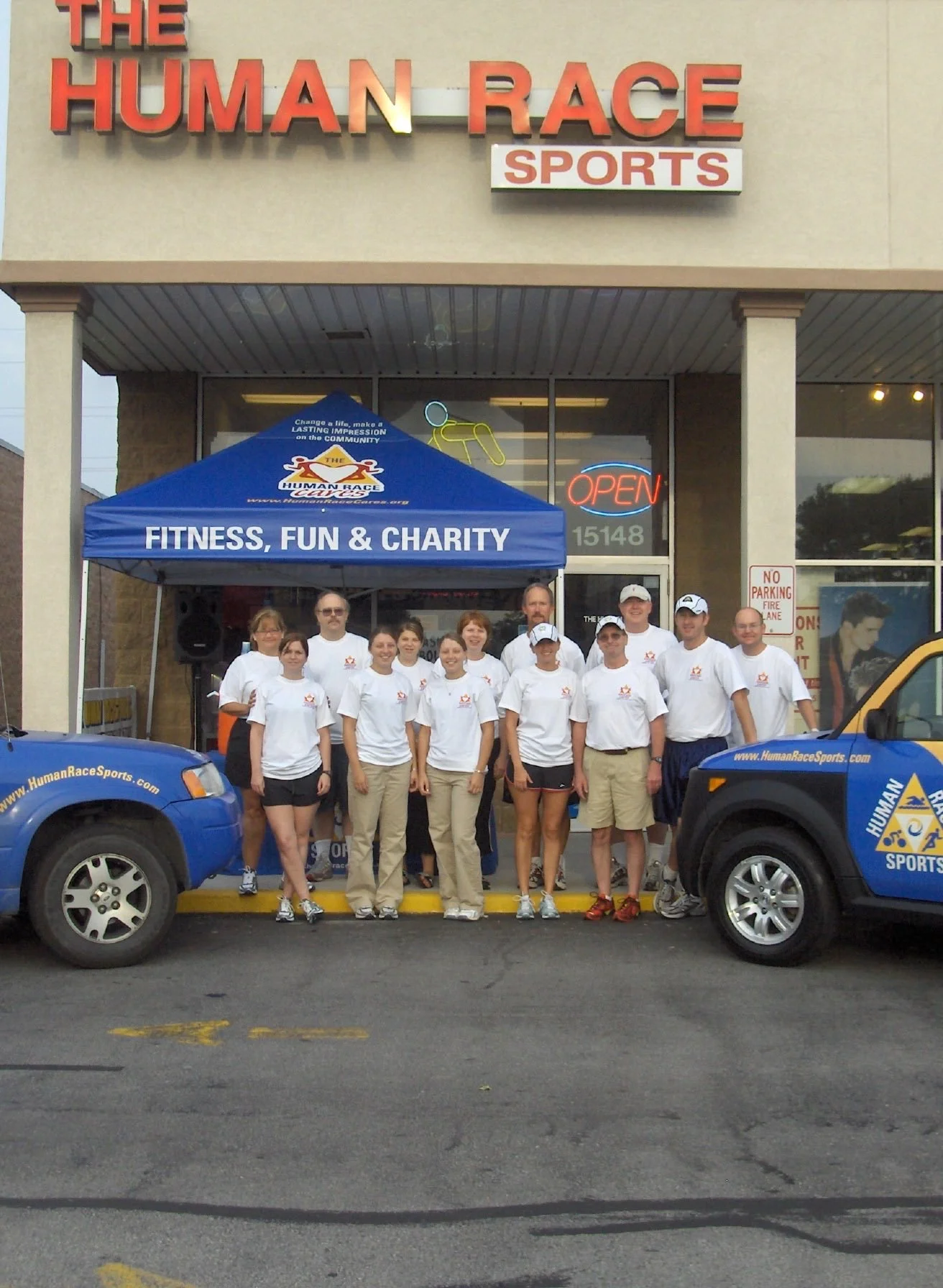 A group of people wearing white shirts standing in front of a building with a sign that says 'The Human Race Sports.' They are under a blue canopy that reads 'Fitness, Fun & Charity.' Two blue vehicles with 'www.HumanRaceSports.com' written on them a