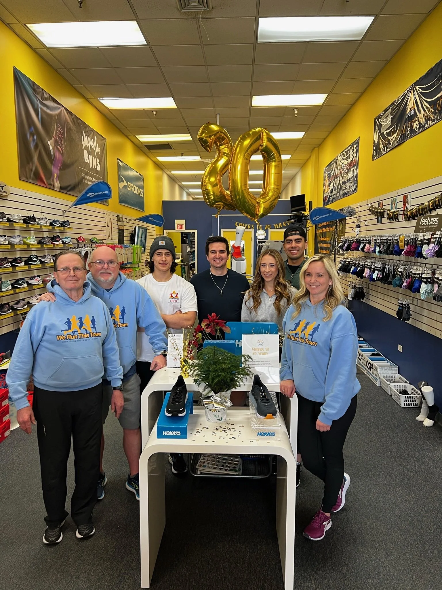 Group of seven people in a sporting goods store celebrating 20th anniversary, with large gold balloons shaped as the number 20, standing behind a display table with running shoes, plants, and decorations.