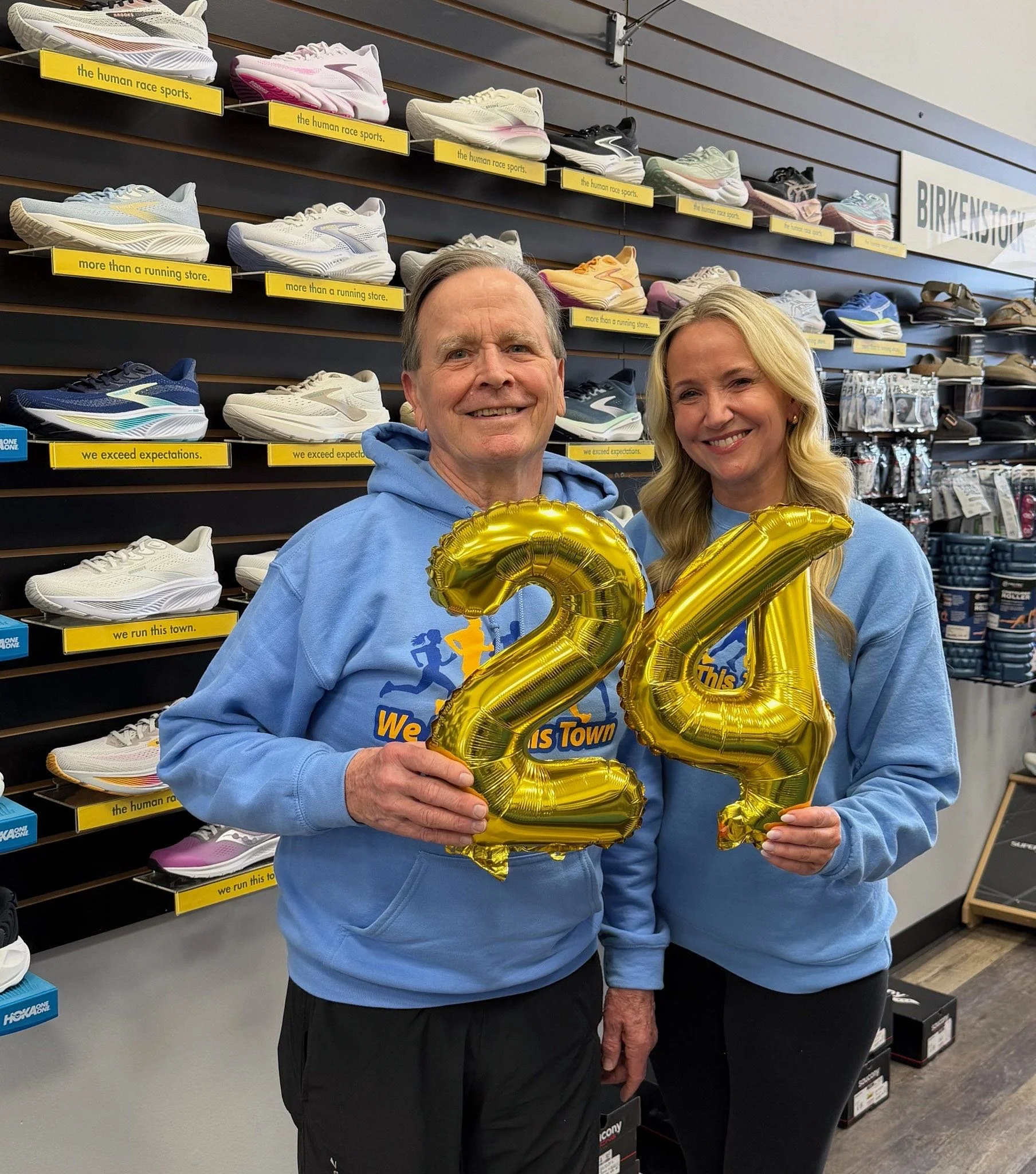 Two people standing inside a store, holding gold balloon numbers '2' and '4', celebrating their 24th anniversary or birthday, with shelves of running shoes in the background.