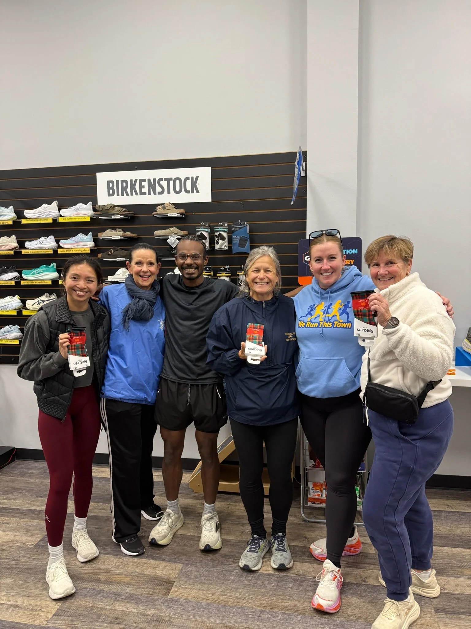 Group of six diverse women and one man standing inside a footwear store, smiling, holding holiday-themed feetures socks, with a wall of shoes behind them and a Birkenstock sign above.
