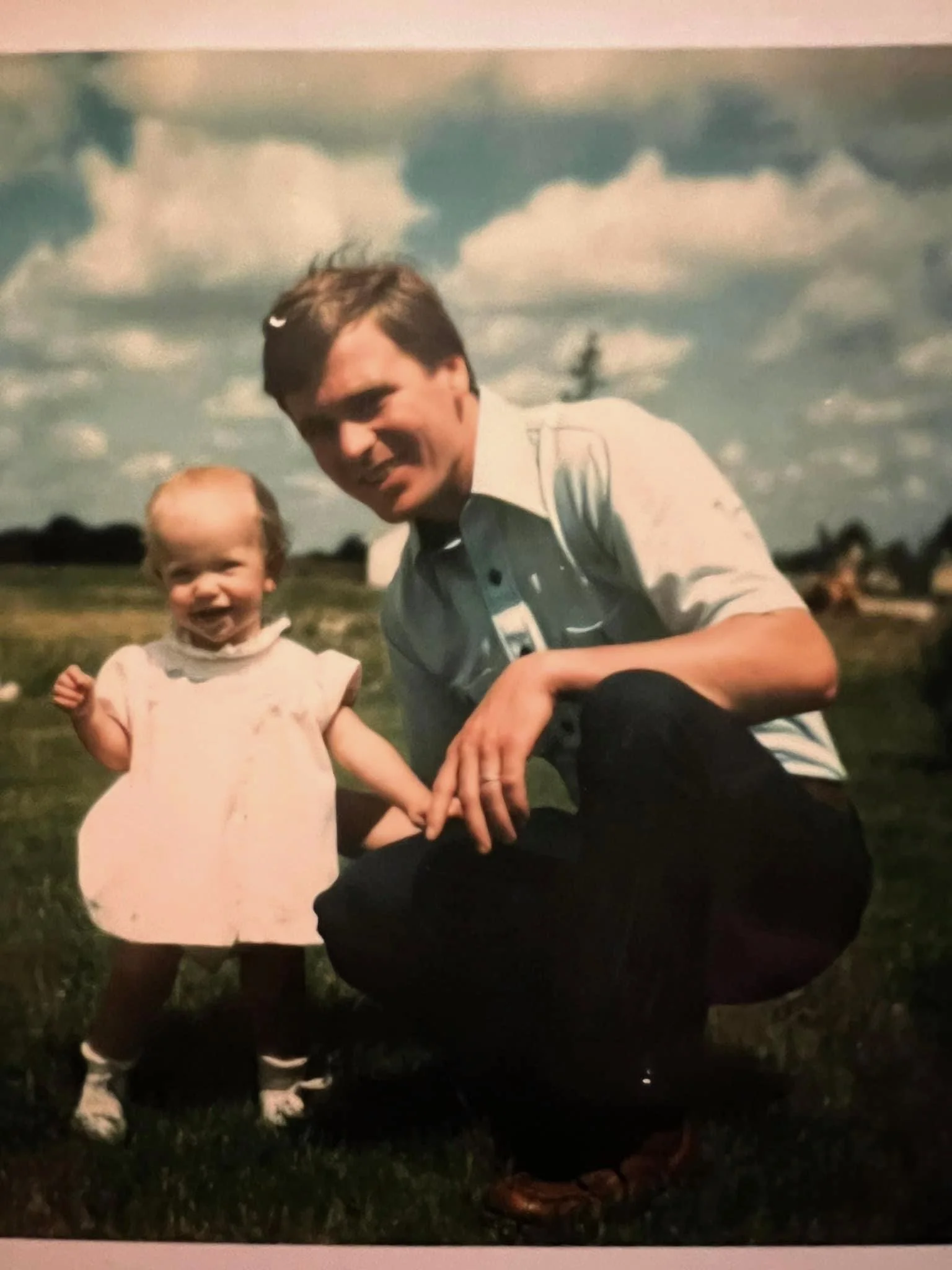 A young man kneeling outdoors on a grassy field, holding the hand of a smiling toddler girl dressed in a light-colored dress and socks, with a partly cloudy sky in the background.