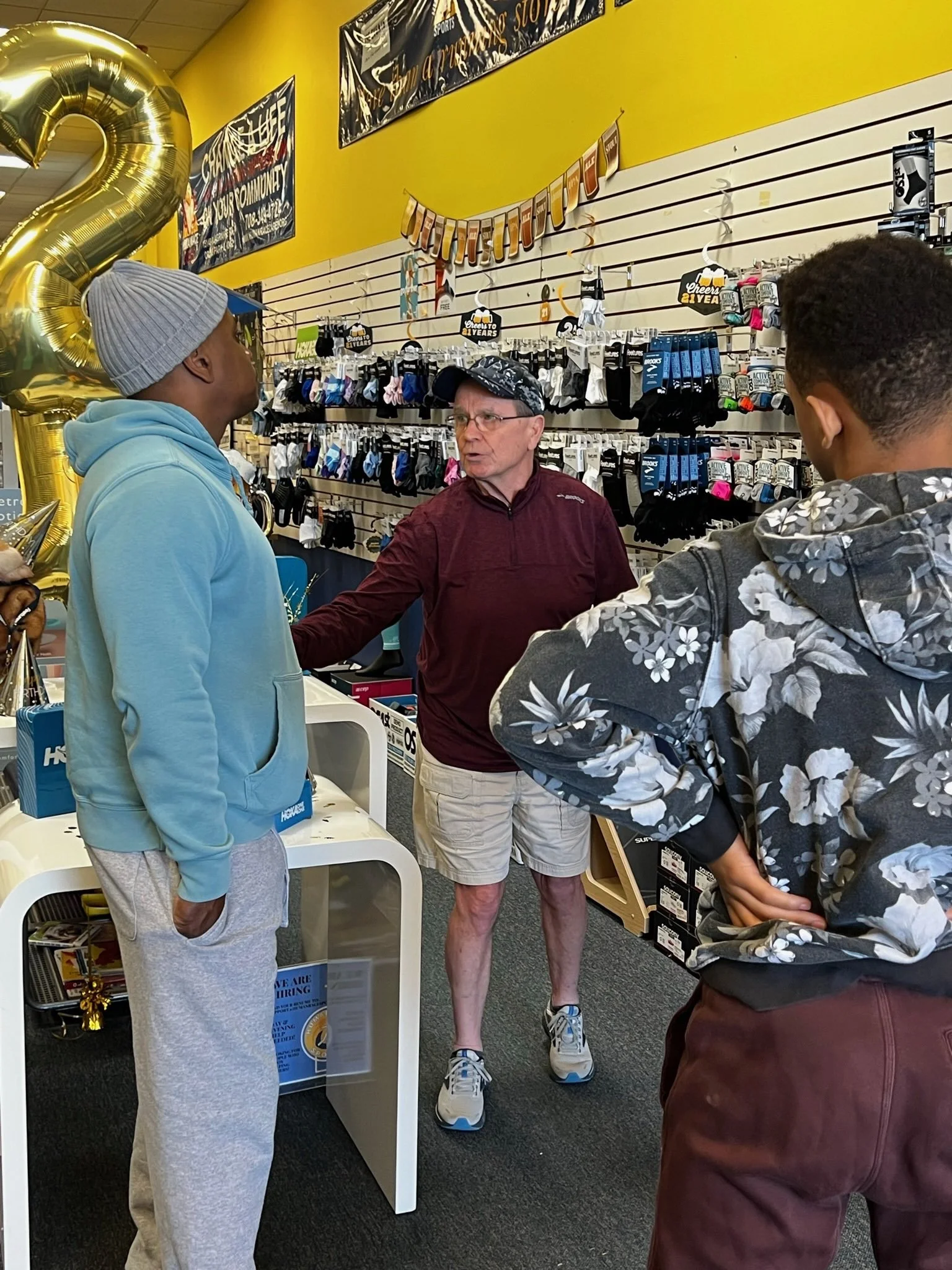 Three people talking inside a store with sporting goods, socks, and underwear on the shelves.