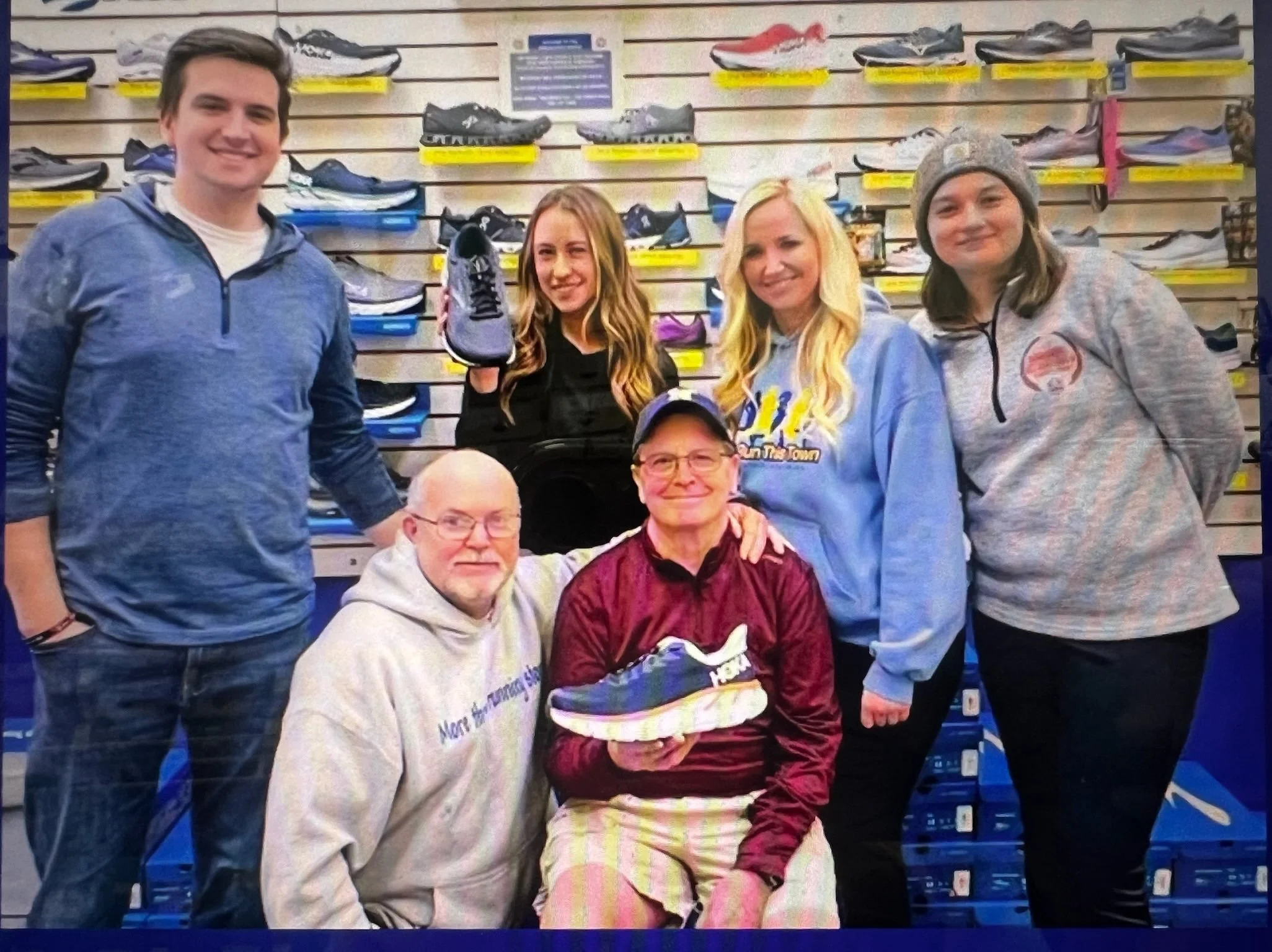 Two men and four women posing together in a shoe store, with shelves of running shoes behind them. One woman holds a running shoe. The group appears happy, and the setting suggests a casual or celebratory moment.