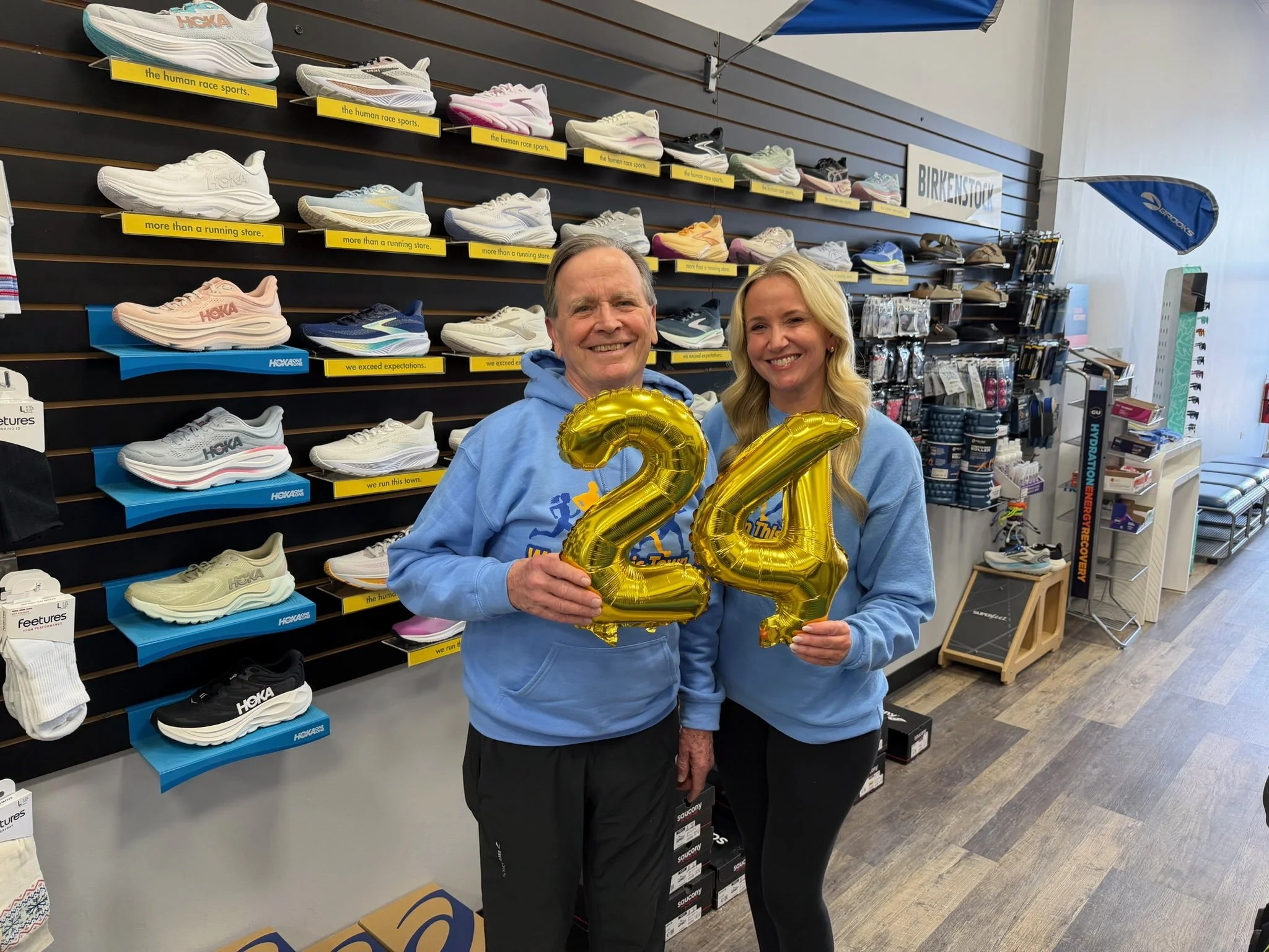 A man and woman standing inside a running shoe store, holding gold balloons shaped as the numbers 2 and 4, celebrating age 24. They are both wearing light blue hoodies and smiling. The store has shelves with various athletic shoes and running accesso