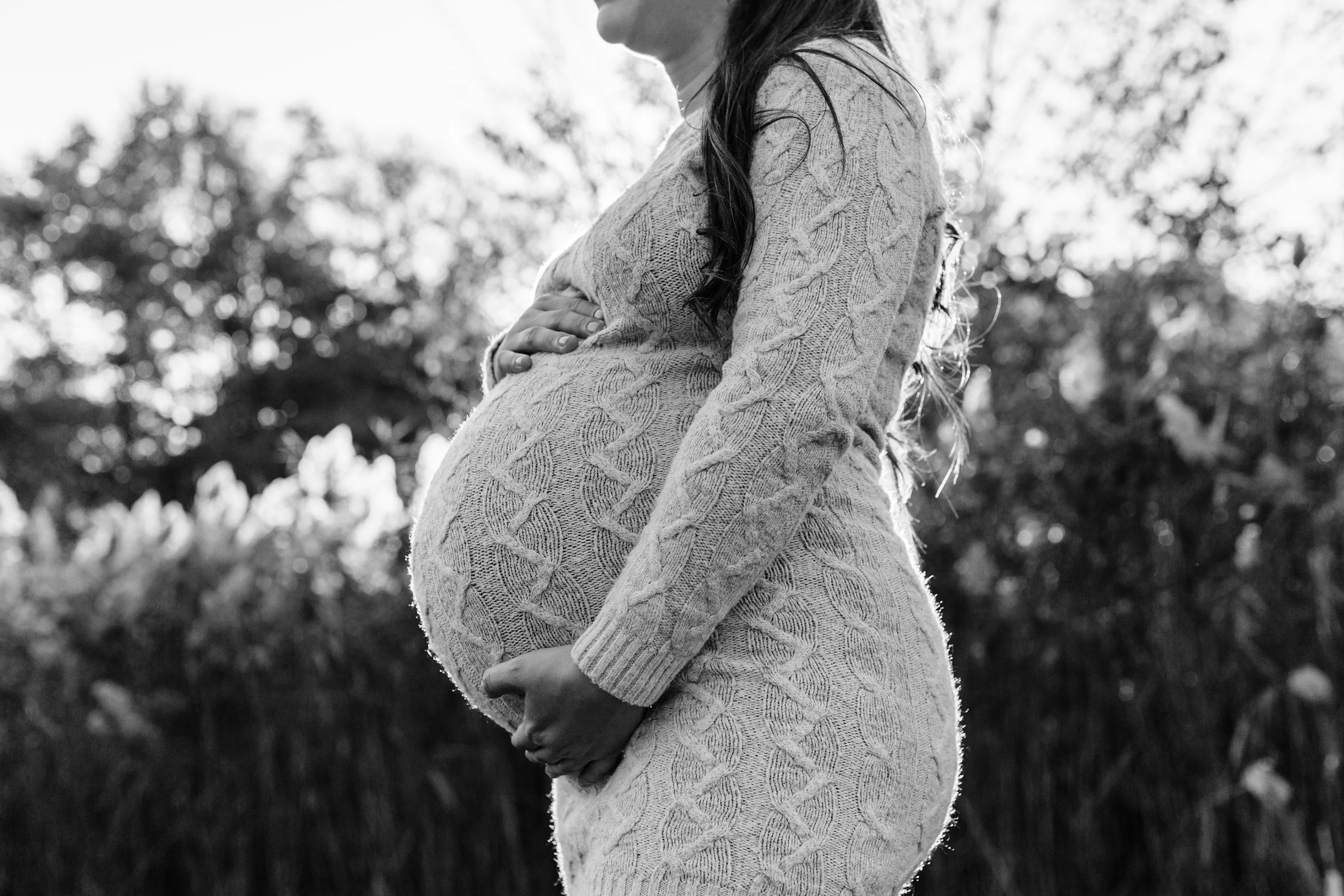 Black and white photo of a pregnant woman in a knitted sweater, holding her belly with her hands, outdoors with trees in the background.