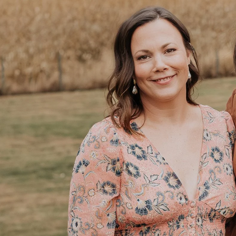 A woman with shoulder-length brown hair smiling outdoors, wearing a pink floral dress and teardrop earrings.