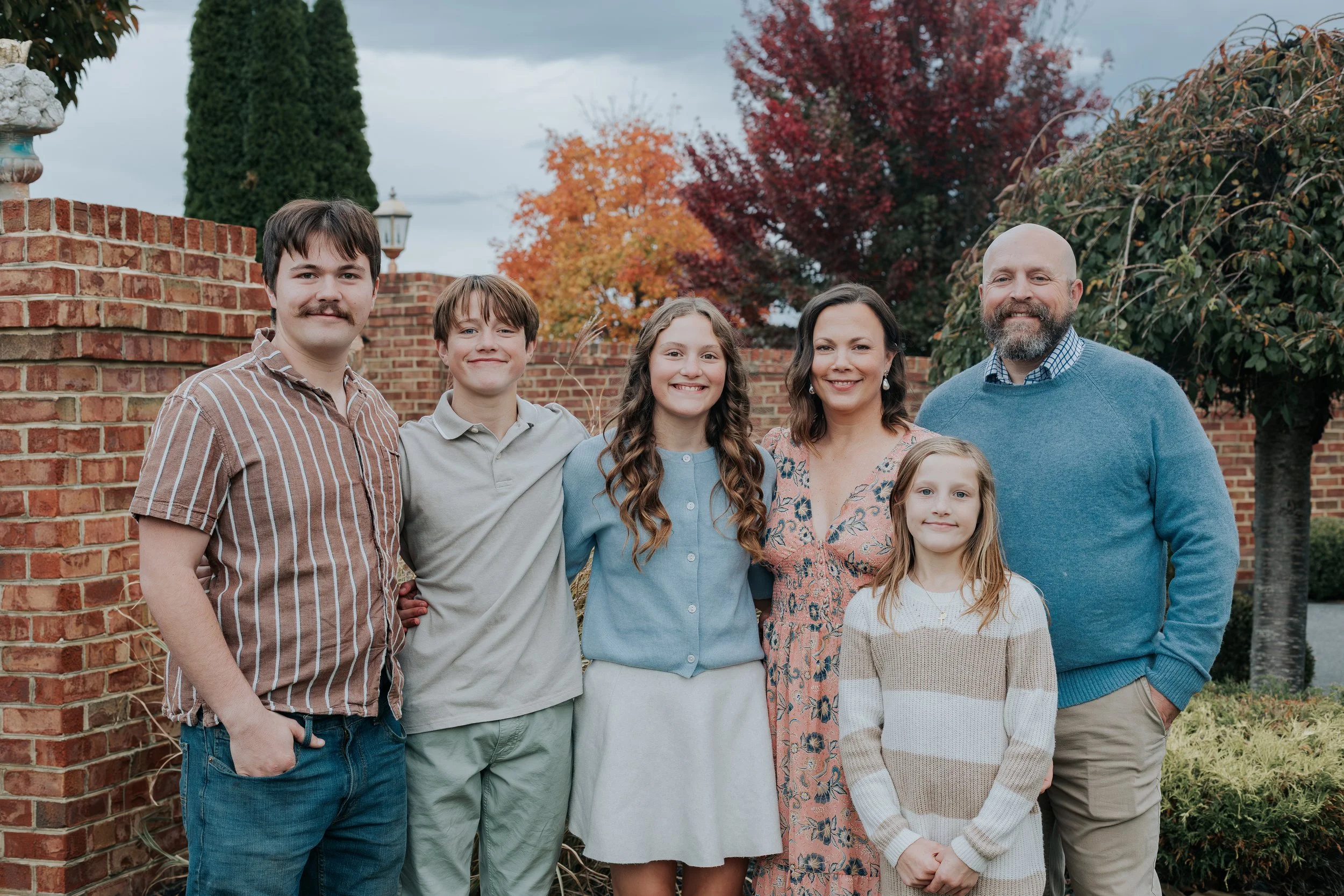 A family of seven standing outdoors during fall, smiling at the camera, with red and orange trees, brick wall, and cloudy sky in the background.