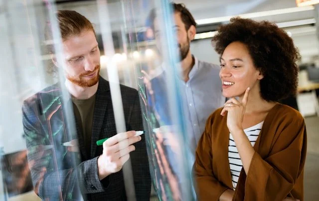 Three colleagues discussing around a transparent board in an office.