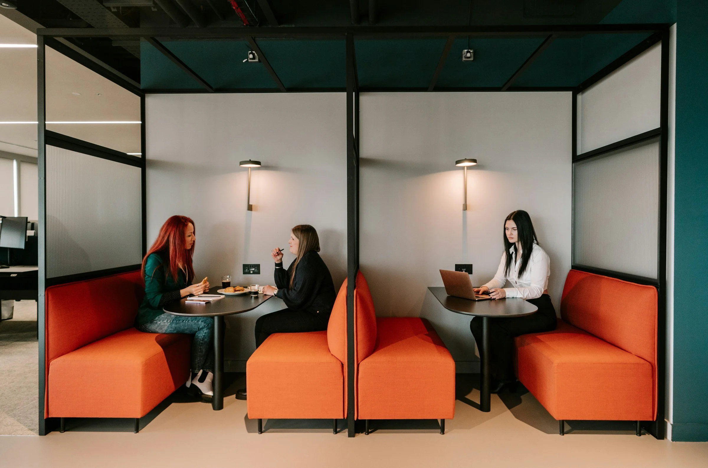 Two women are sitting at separate booth-style small tables with orange cushioned seating in an office setting. The woman on the left has red hair and is wearing a dark green top, holding a snack. The woman on the right has brown hair and is dressed in black, also eating. On the right side, another woman with black hair, dressed in white, is working on a laptop at a similar booth.