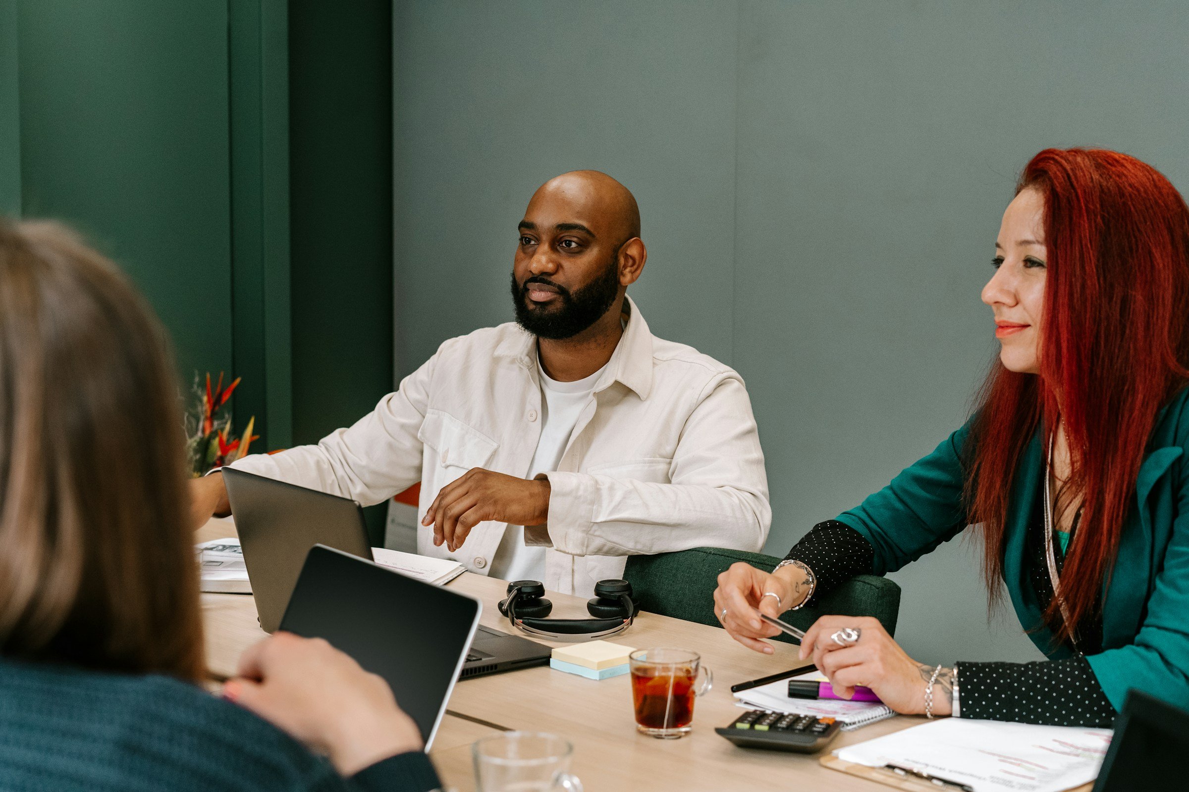 A diverse group of four people having a meeting at a conference table in an office. One man with a beard and a white shirt is speaking, while a woman with red hair, green blazer, and polka-dot cuffs is listening, holding a pen. Two laptops, notepads, a calculator, headphones, and a glass of tea are on the table.