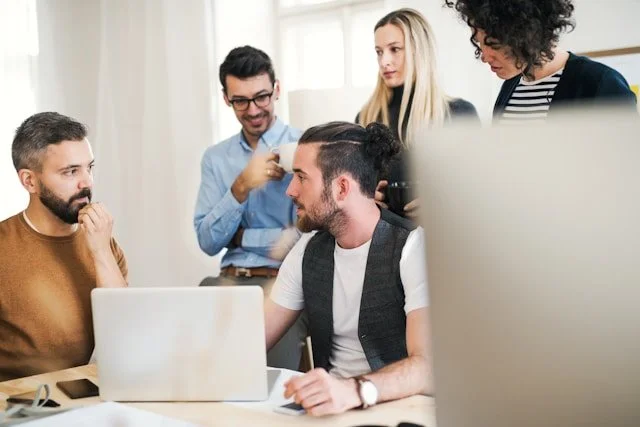 Group of five young adults in a meeting room, engaged in discussion around a laptop.