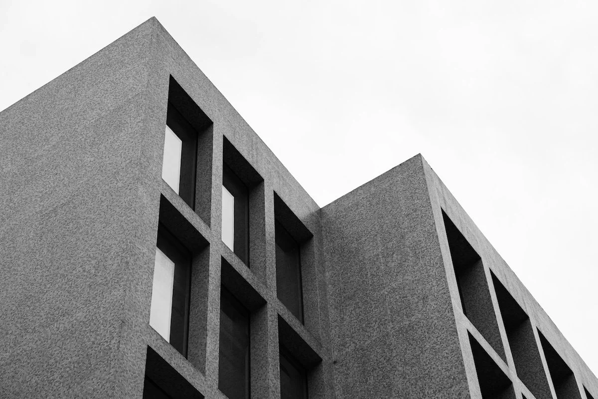 Black and white photo of a modern building with a textured facade and rectangular windows, viewed from an angle against a cloudy sky.
