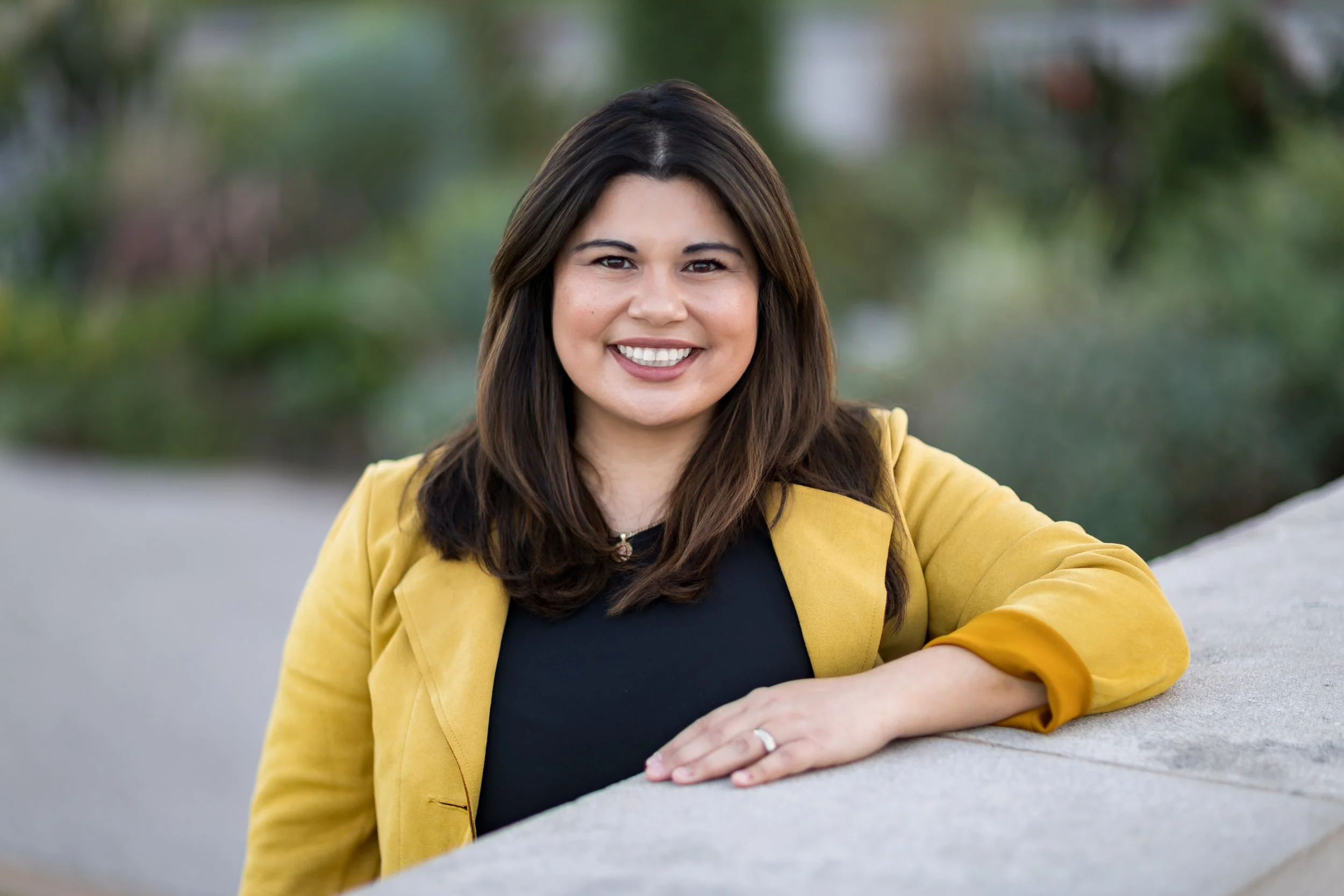 A woman with shoulder-length dark hair, wearing a yellow blazer over a black top, smiling outdoors with blurred greenery in the background.