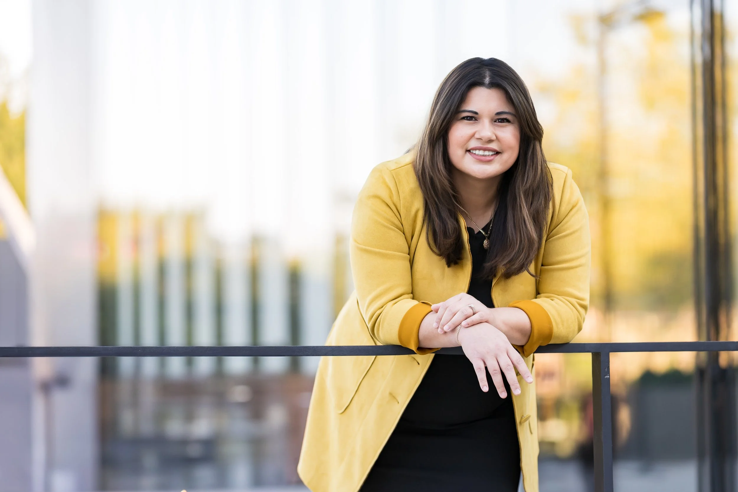 A woman with long brown hair in a yellow blazer leaning on a black railing outdoors, with a blurred background of trees with fall foliage.