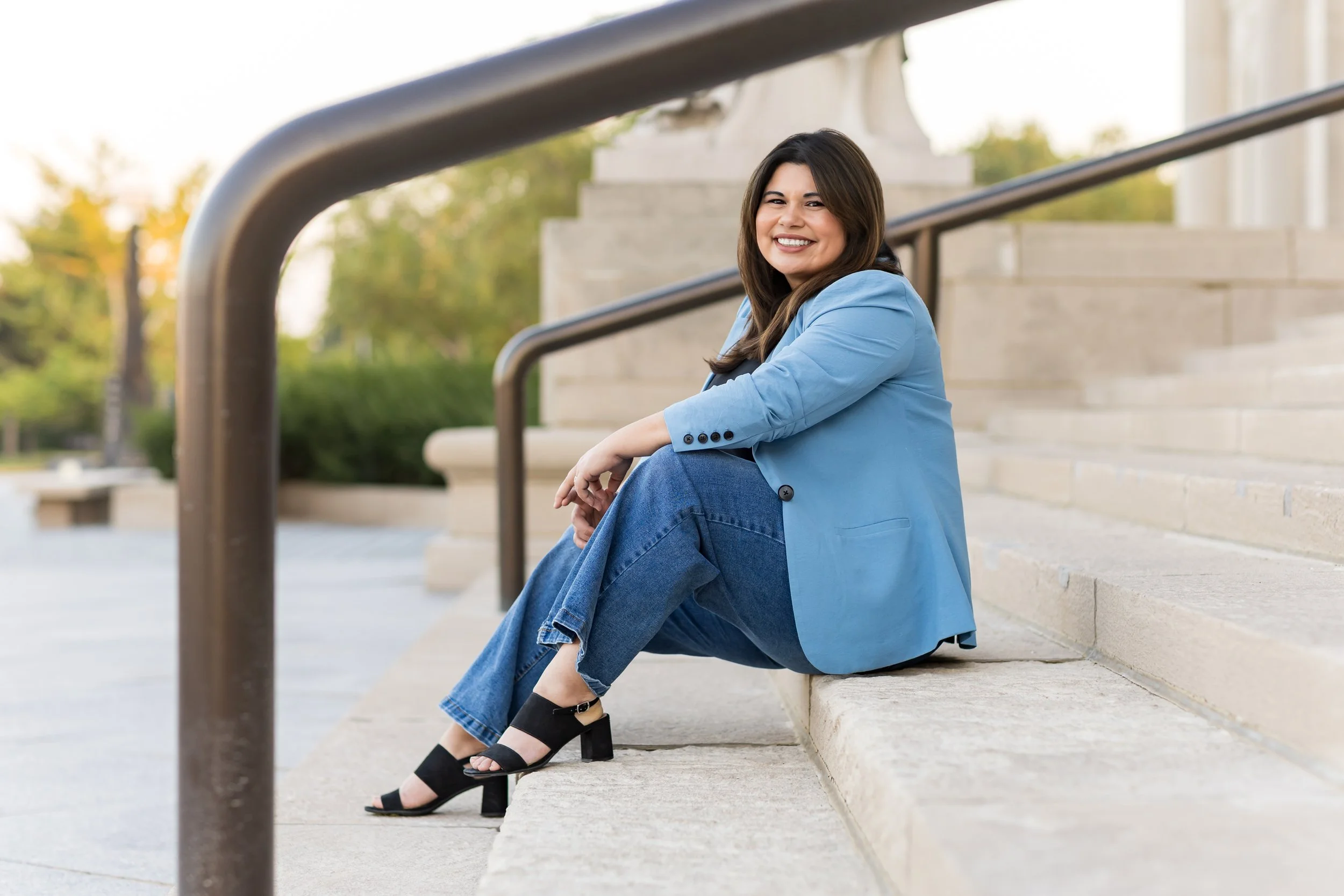 Woman sitting on steps outdoors, smiling, wearing a blue blazer, jeans, and black heels, with a statue and trees in the background.