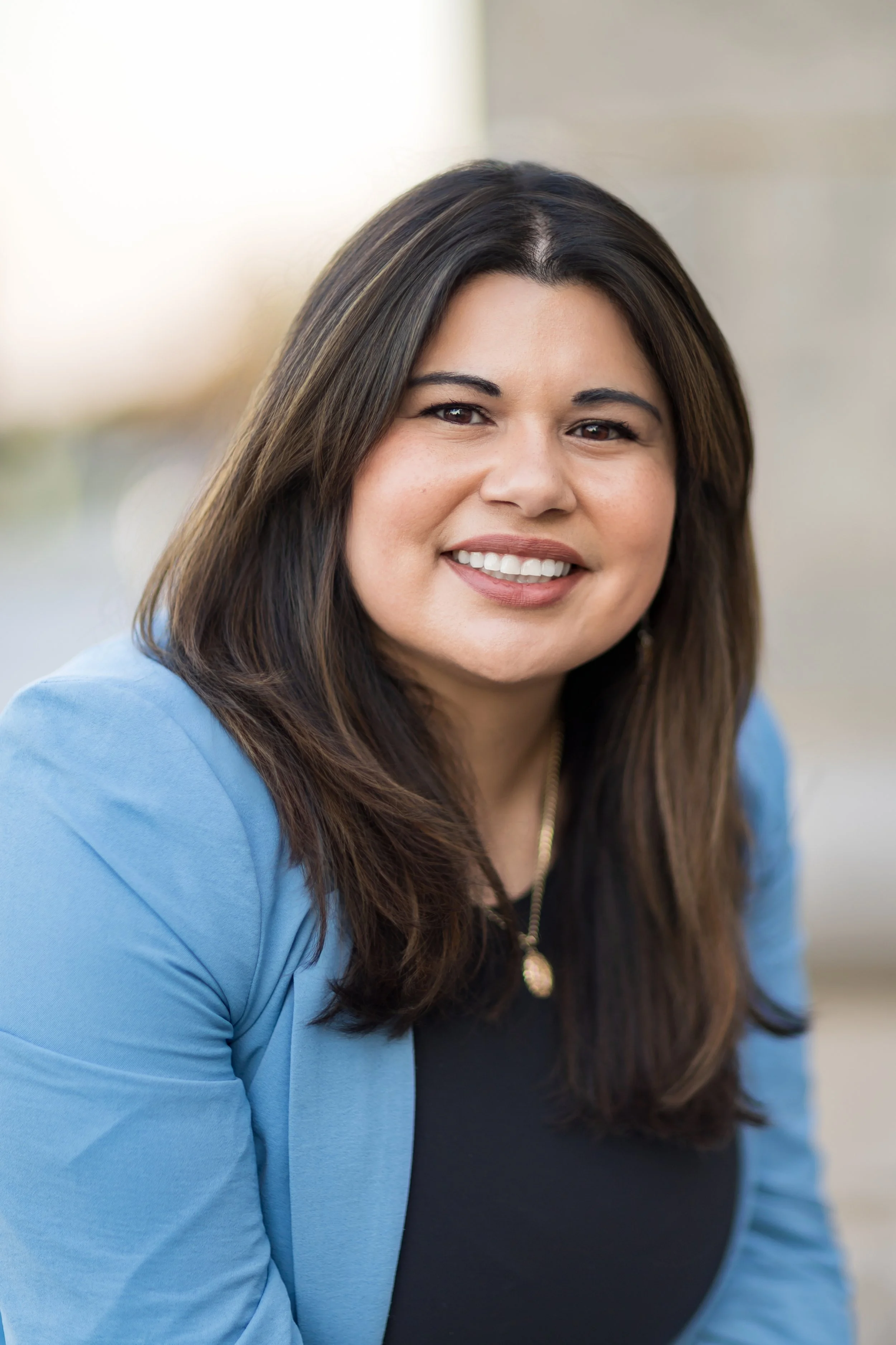 A woman with long brown hair smiling, wearing a blue blazer and black top, outdoors with a blurred background.