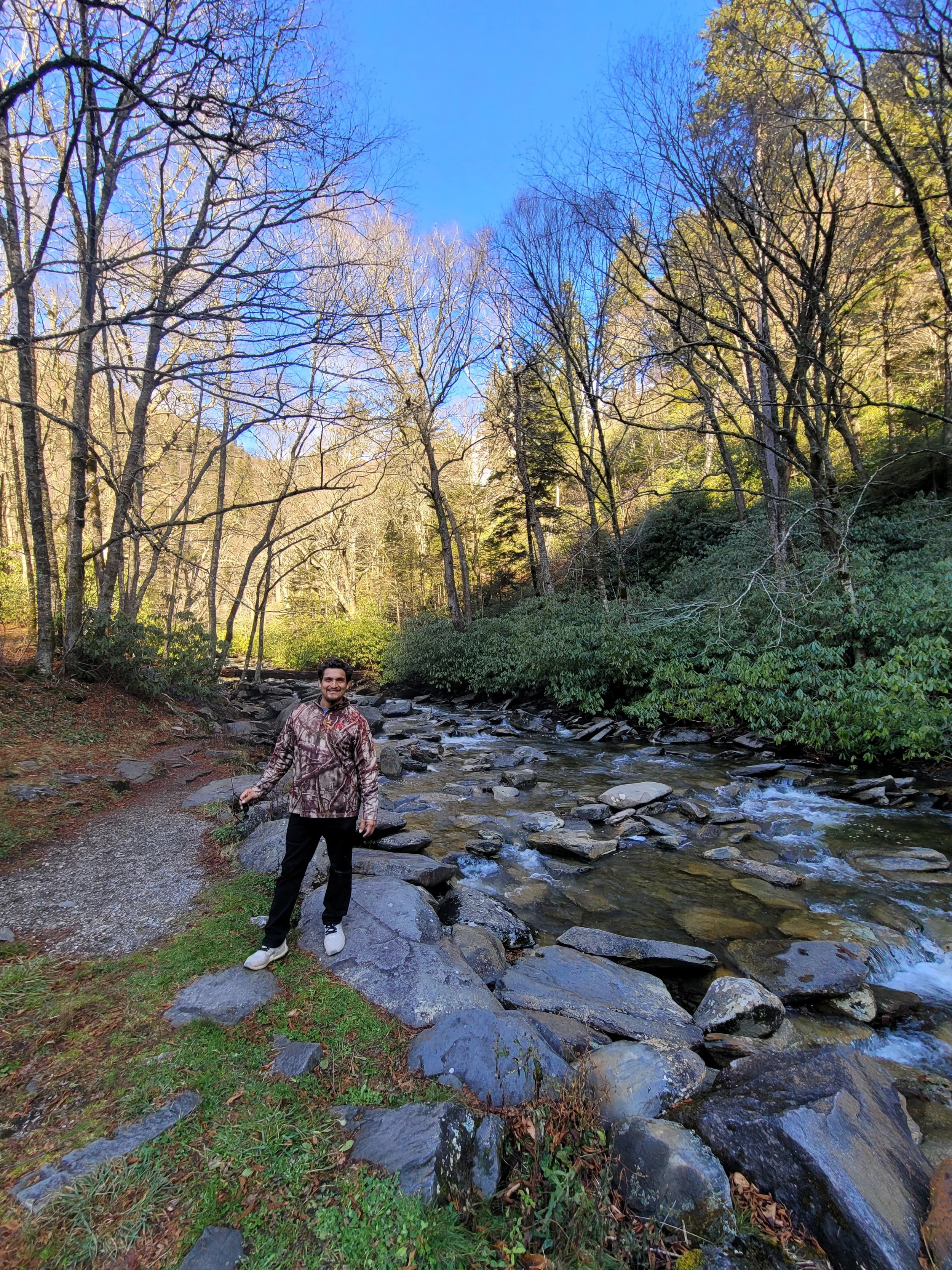 An arborist smiling standing on rocks by a creek in a forested area with leafless trees and some green shrubs, under a partly cloudy blue sky.