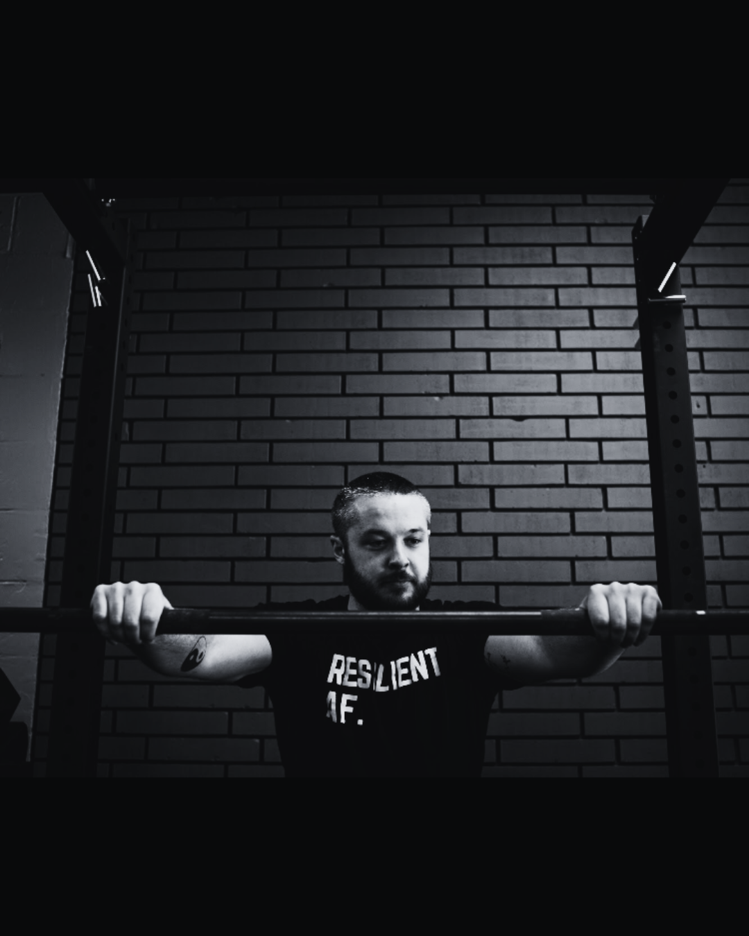 A man lifting a barbell in a gym with a brick wall background, black and white photograph.