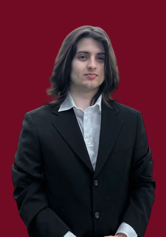 Young man with long dark hair wearing a black suit and white shirt standing against a solid red background.