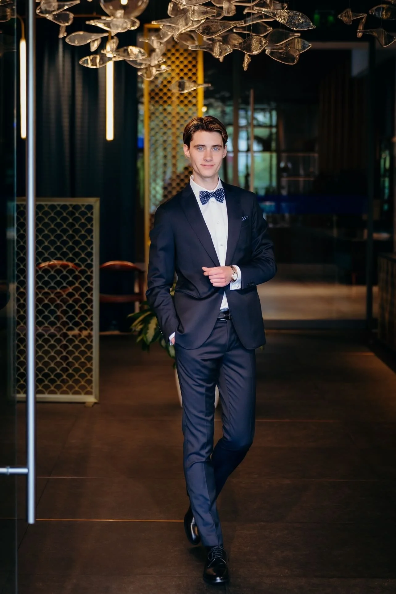 A young man in a dark suit with a white shirt and a polka dot bow tie, standing in a stylish, modern interior with decorative hanging glass art and warm lighting.
