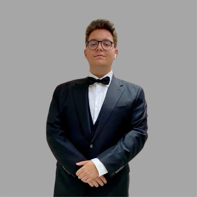 A young man wearing glasses, a black tuxedo with a bow tie, and white dress shirt, standing against a plain gray background with a confident expression.